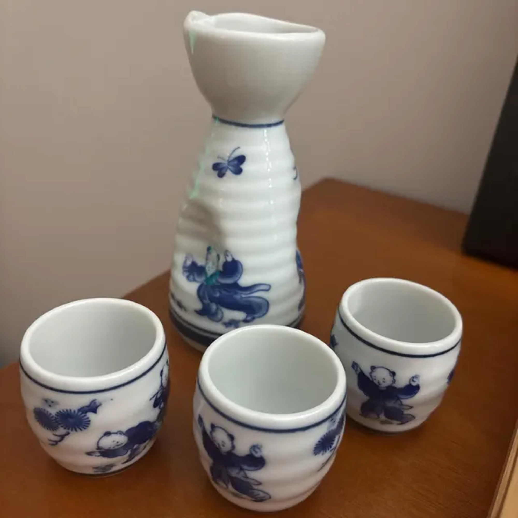 Set of seven white ceramic sake cups with blue floral and butterfly patterns, arranged on a wooden table.