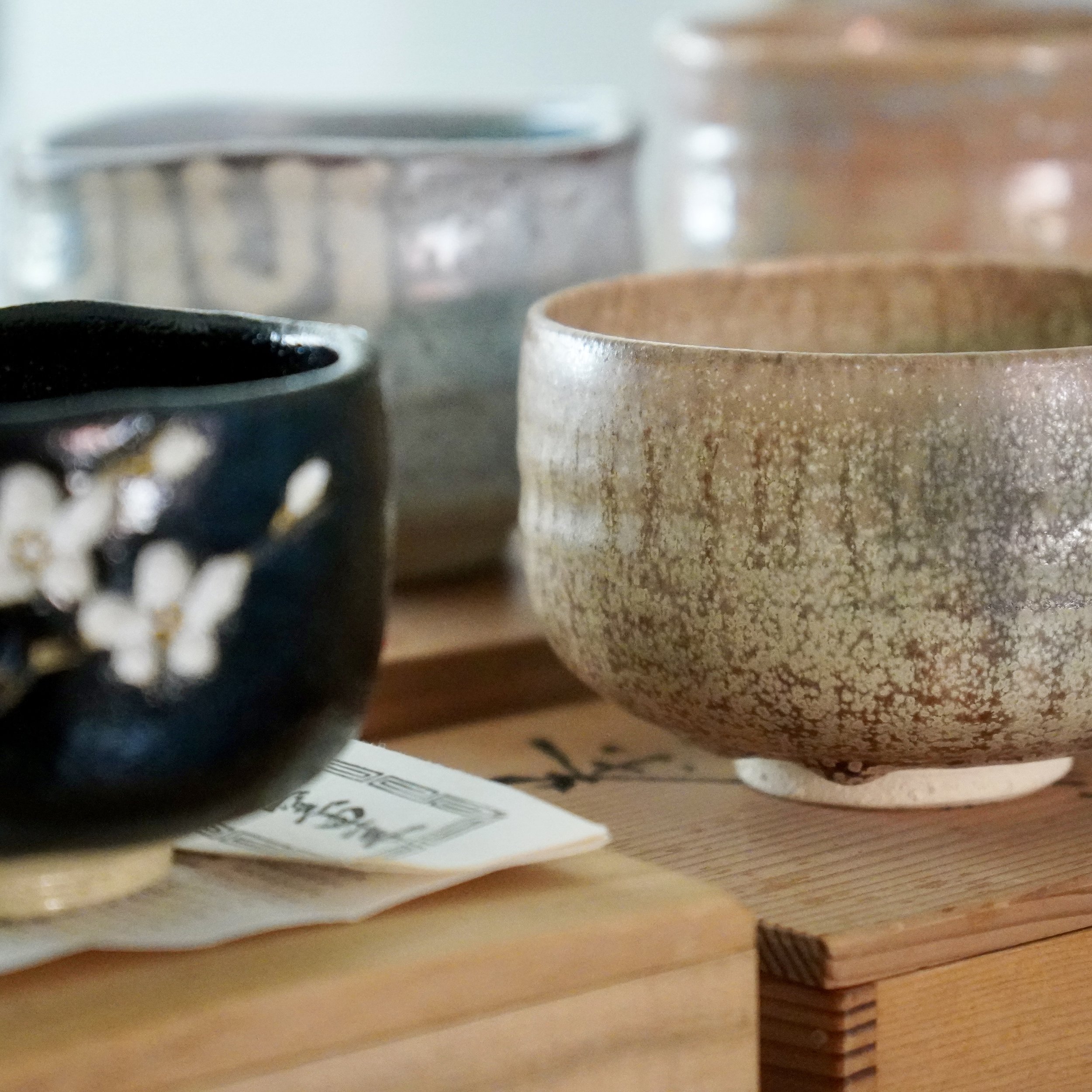 Japanese matcha bowls on a wooden surface: one with a dark iron glaze and cherry blossom detail, one in rough natural stoneware. Vintage chawan for the Japanese tea ceremony, sourced in Japan.