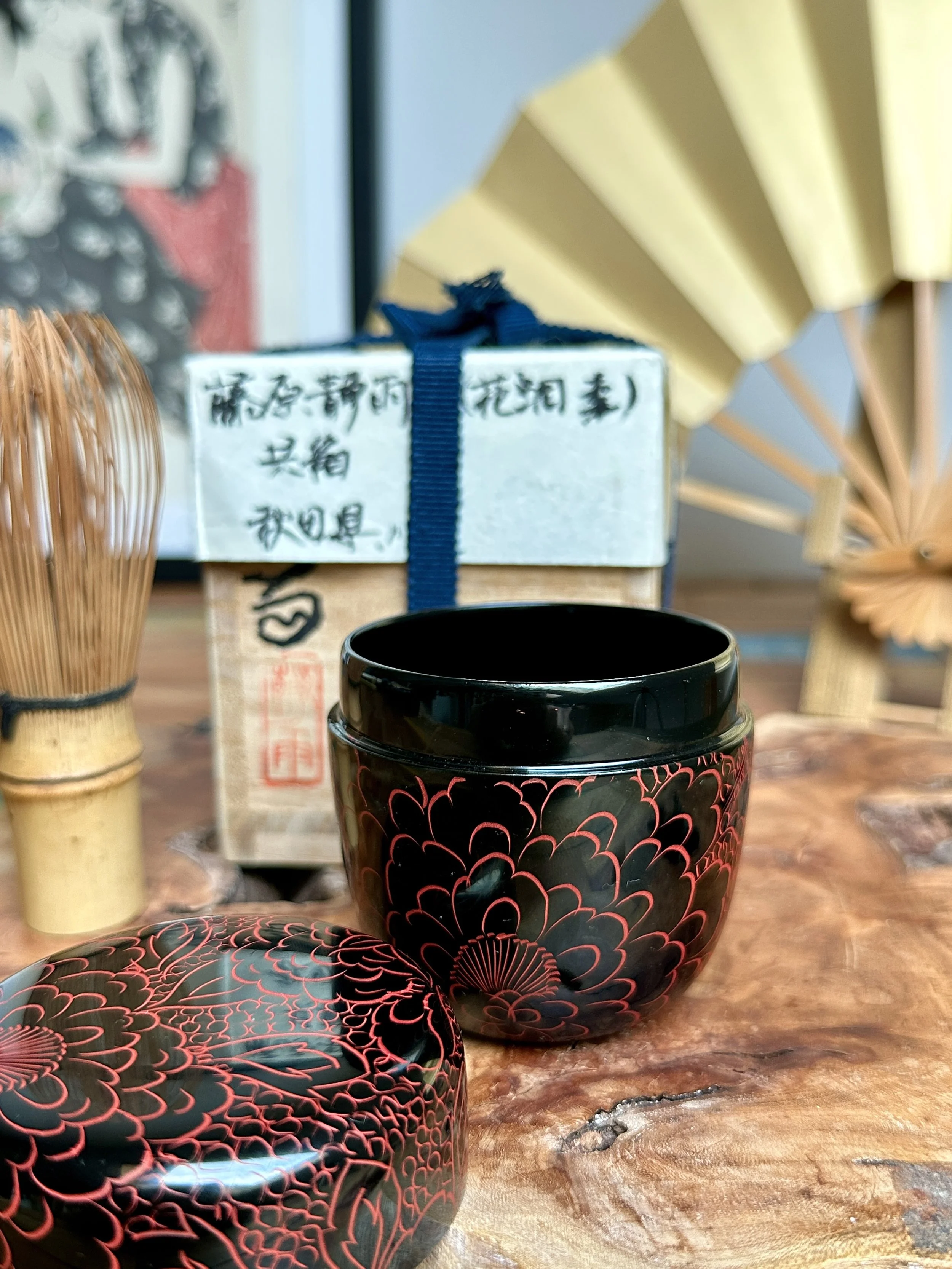 Black lacquered tea caddy by Fujiwara Seiu with red peony design, open lid, matcha whisk, and tomobako box on a wooden surface.