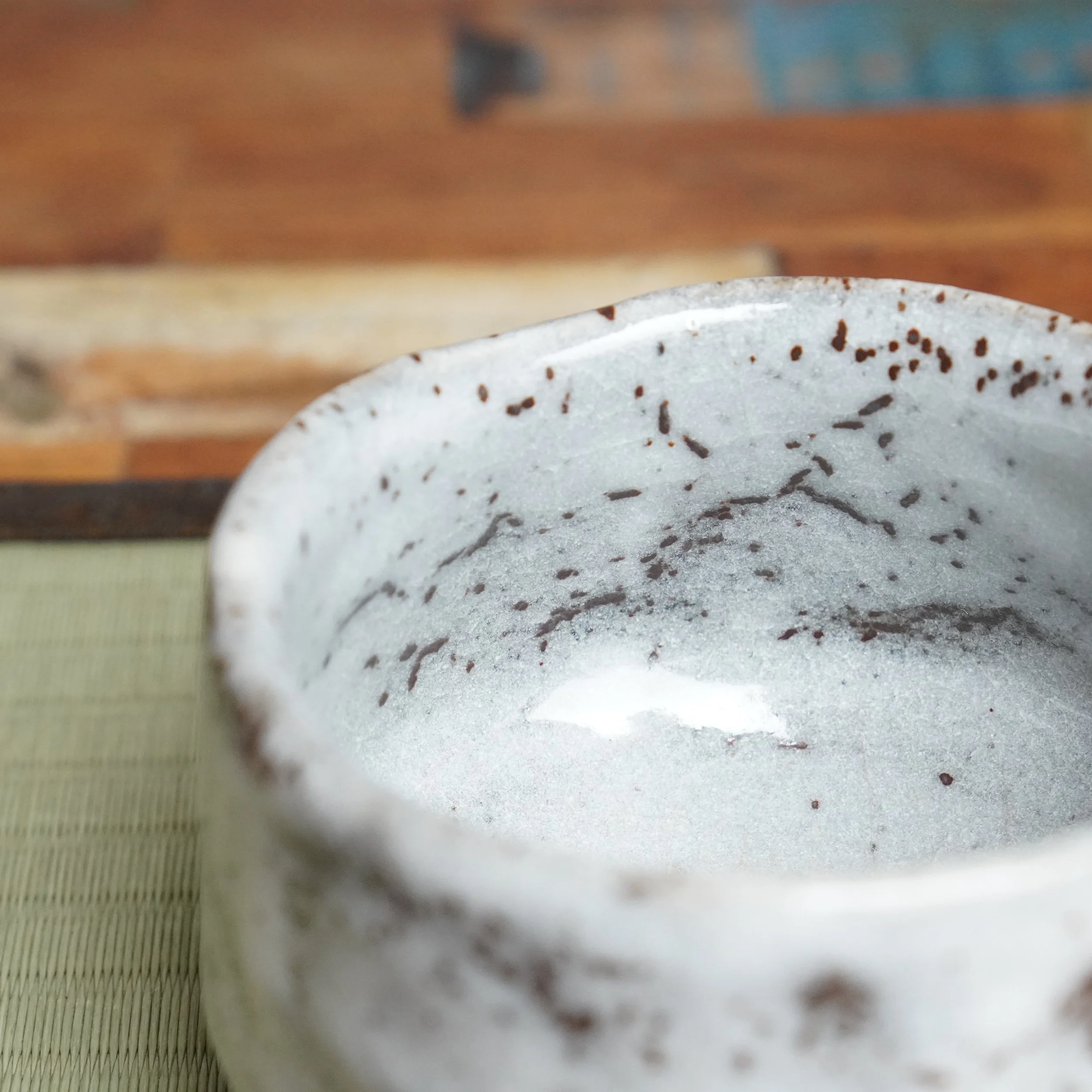 Close-up view of a ceramic tea bowl by Katō Shōzan, with a thick, speckled white glaze and rust brown flecks. The interior reflects light. Blurred wood and a tatami mat are in the background.