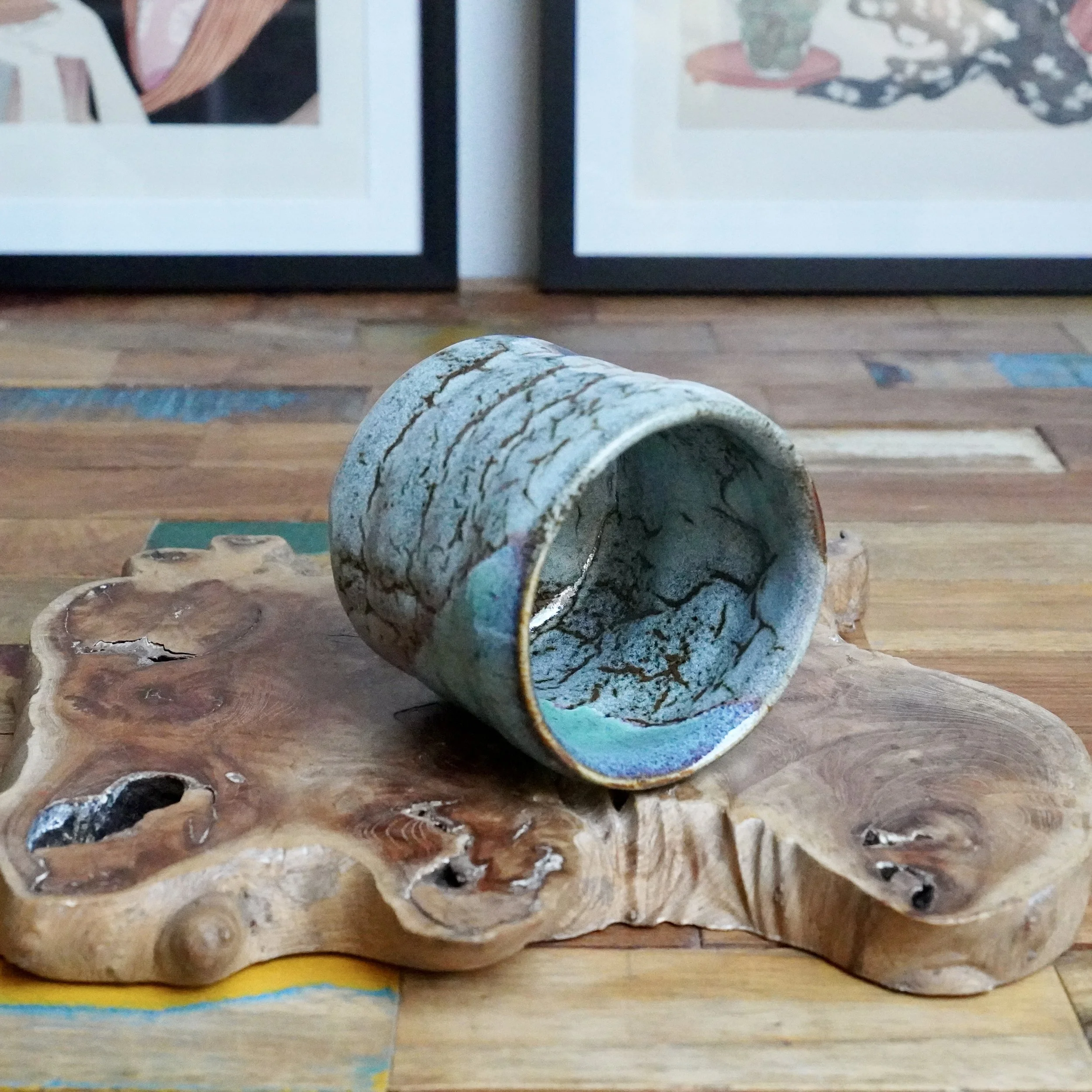 Tsutsu chawan with layered crackled turquoise, grey, and brown glaze, resting on its side on a natural wood slab.