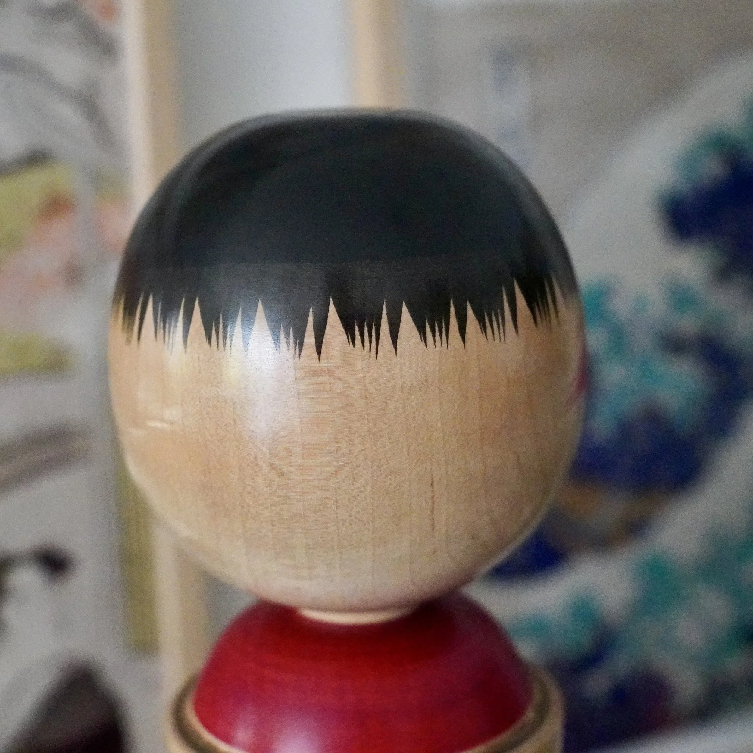 Kokeshi doll head by Izu Mamoru, pale wood, with a precise black bob and triangular fringe, above a red body.