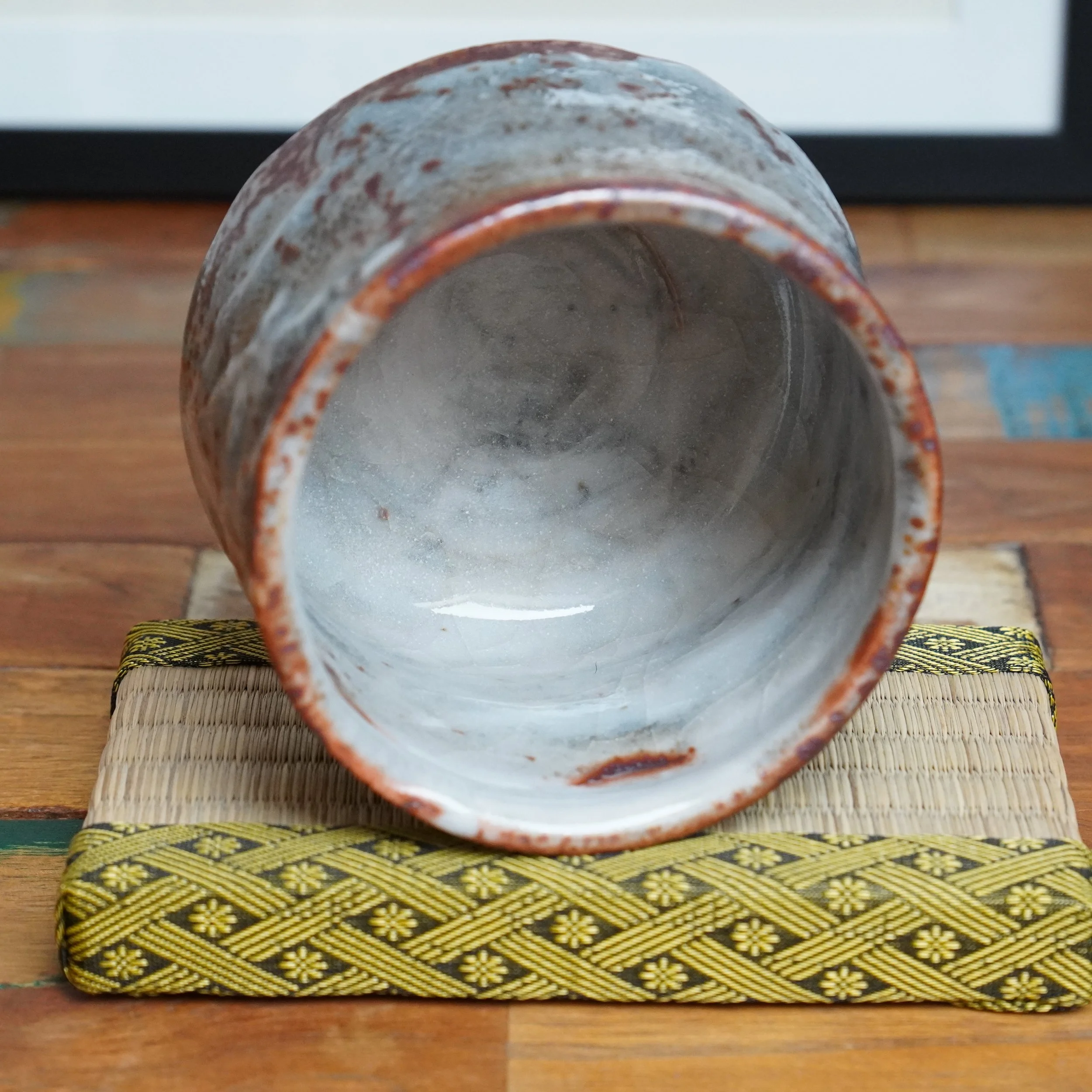 Shino chawan, on its side, displays its cracked white interior and grey-white exterior with reddish-brown speckles. It rests on a woven mat.