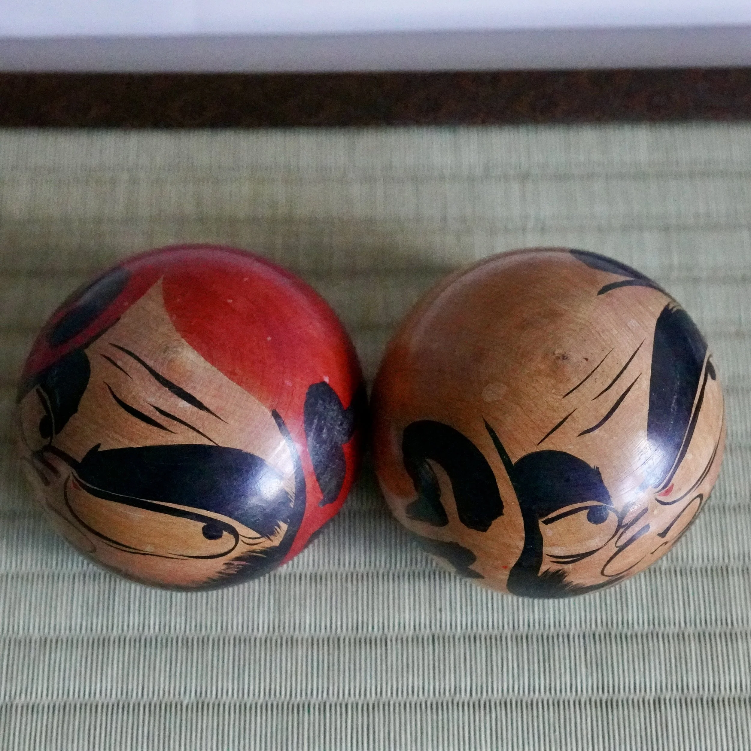 Two round Tsugaru daruma by Satō Zenji, Nuruyu. One red-painted, one natural wood, both with black stylized scowling faces on a woven mat.