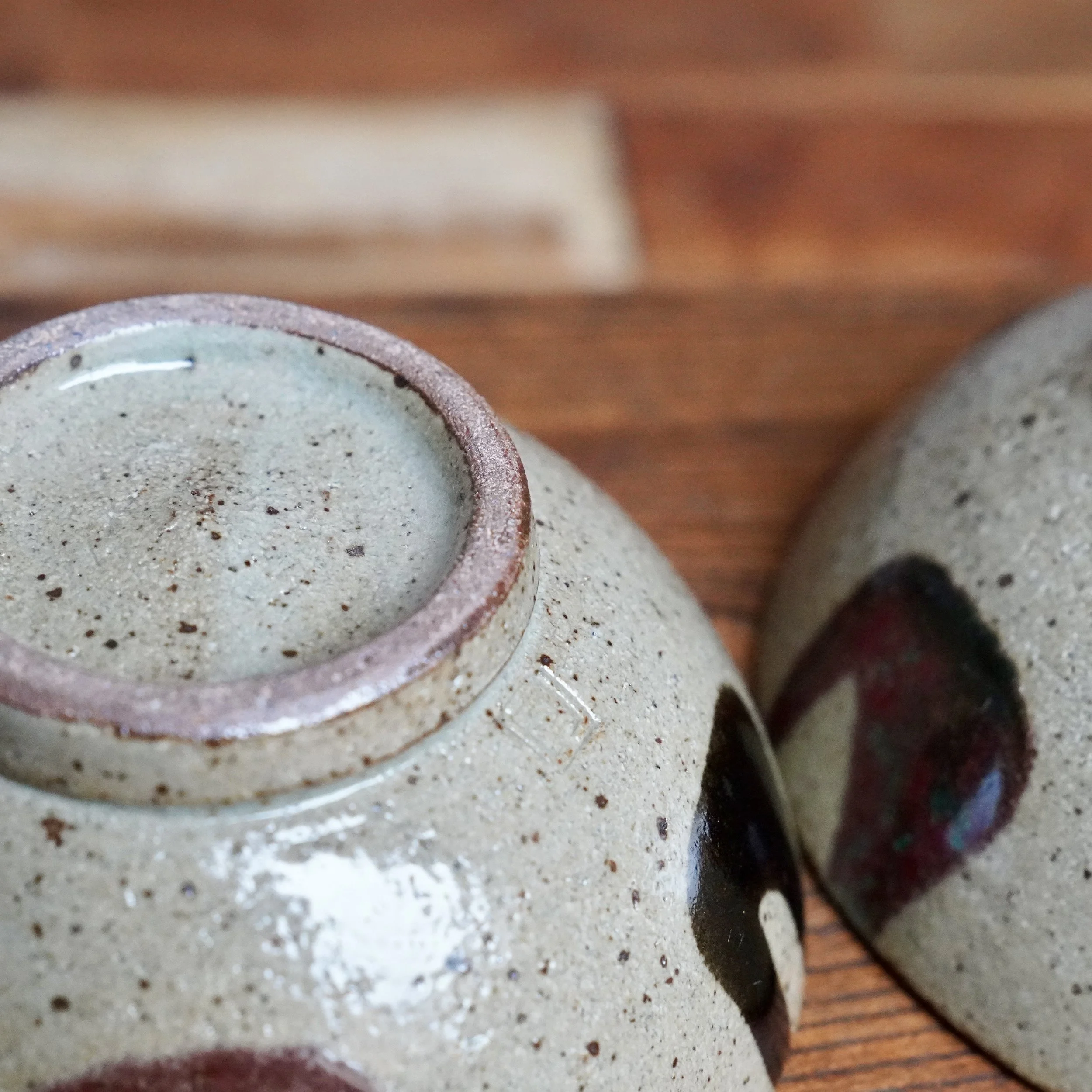 Undersides of two ceramic tea bowls with pale grey speckled clay, reddish foot rims, and shiny pale green-grey, near-black, and wine-red glazes.