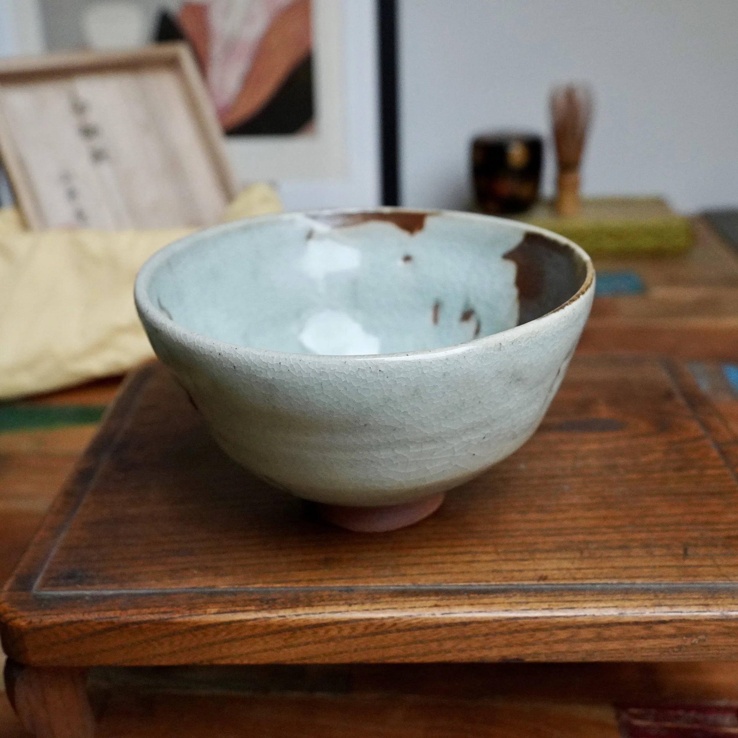 Sansōan chawan with pale blue-grey ice crackle and a deep rust-brown flowing glaze, showing its reddish-brown foot. It rests on a wooden tray with a blurred tomobako and whisk.