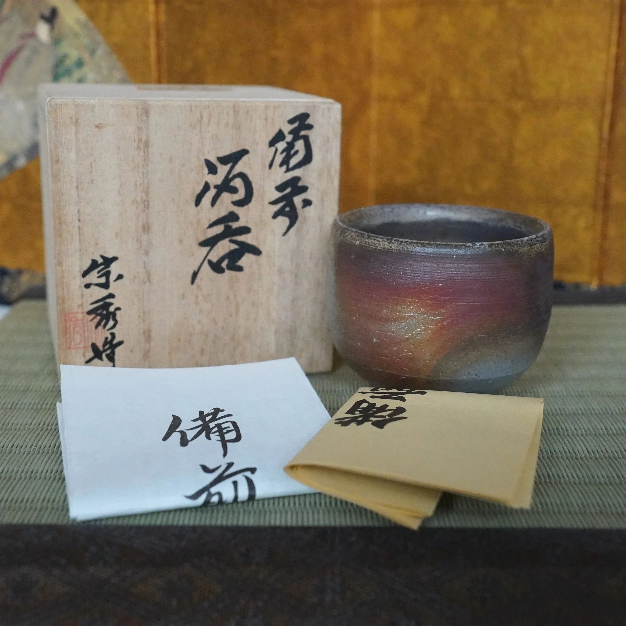 Bizen-yaki guinomi with aubergine, red-gold, and grey sangiri marks. Next to it, a wooden box featuring Yamamoto Sōshū's calligraphy and red seal, plus a white and a tan folded paper.