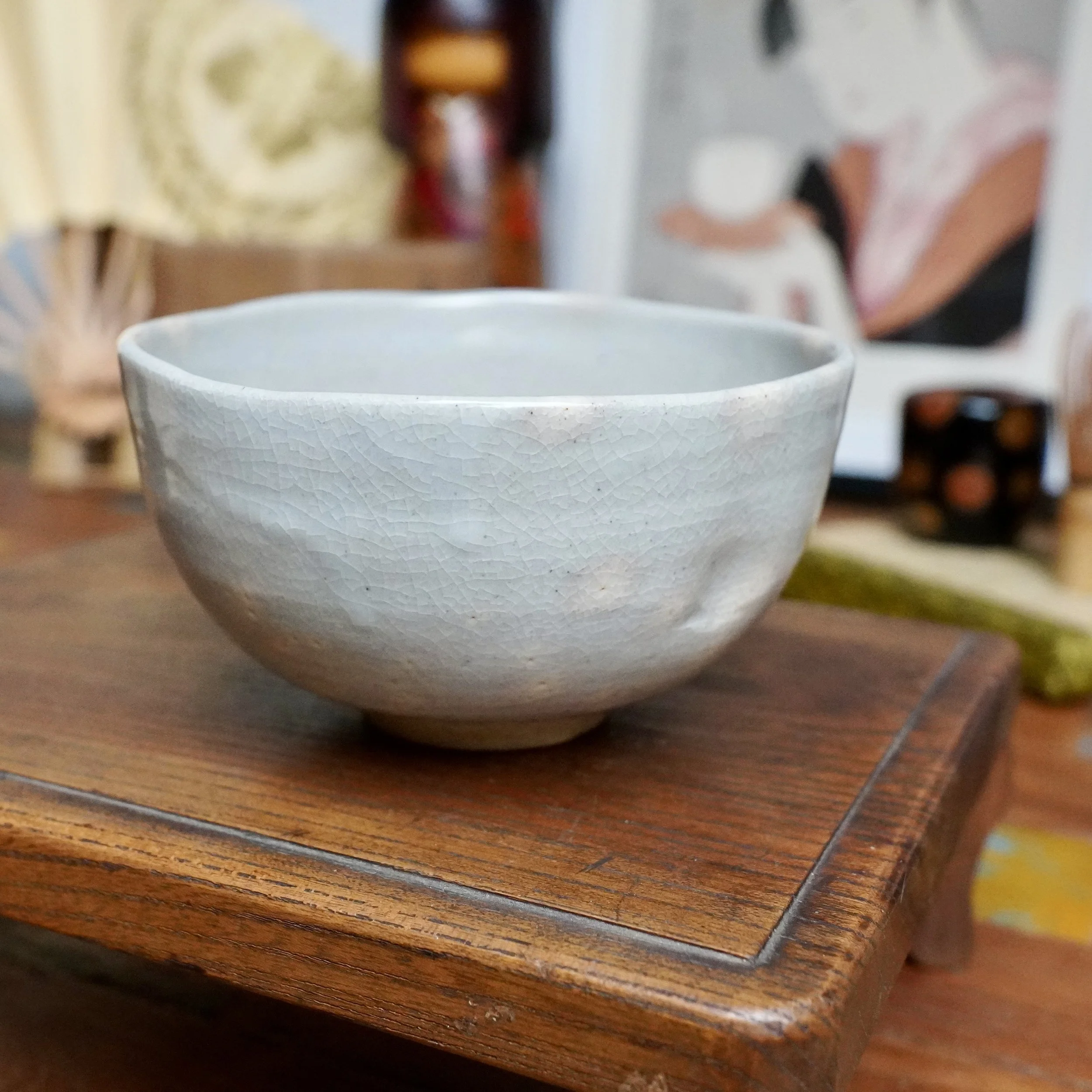 Nagasawa Minoru chawan with a soft grey-blue crackled glaze and faint pink blush. One side of the rim is gently pressed, resting on a wooden stand.