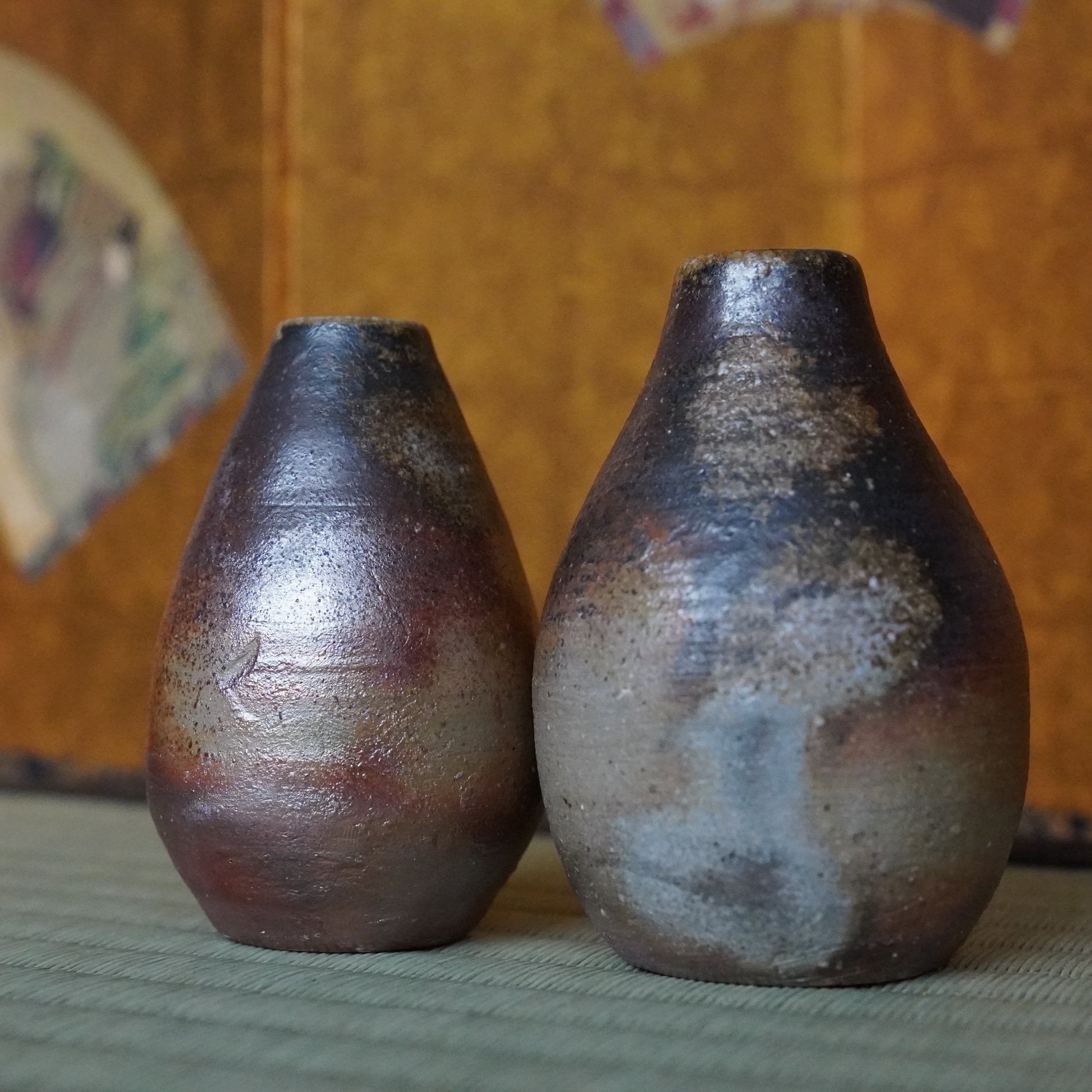 Shibaoka Tōsendō, Kōzan-gama Bizen-yaki tokkuri pair on tatami. One dark aubergine with silver, the other silvery-grey with rust tones.