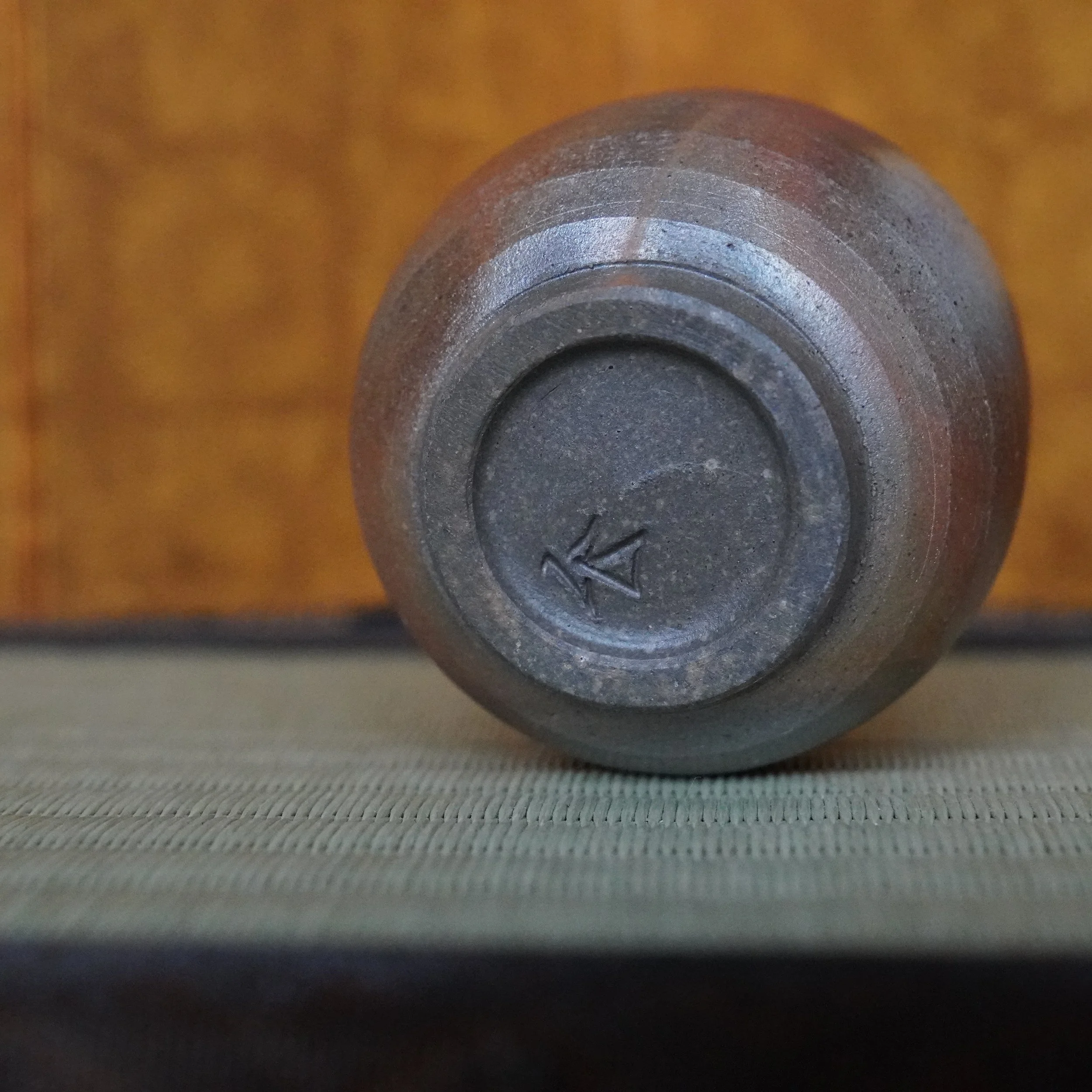 Underside of a dark grey Bizen-yaki guinomi, featuring the foot ring with a carved Yamamoto Sōshū signature, resting on a light green tatami mat.