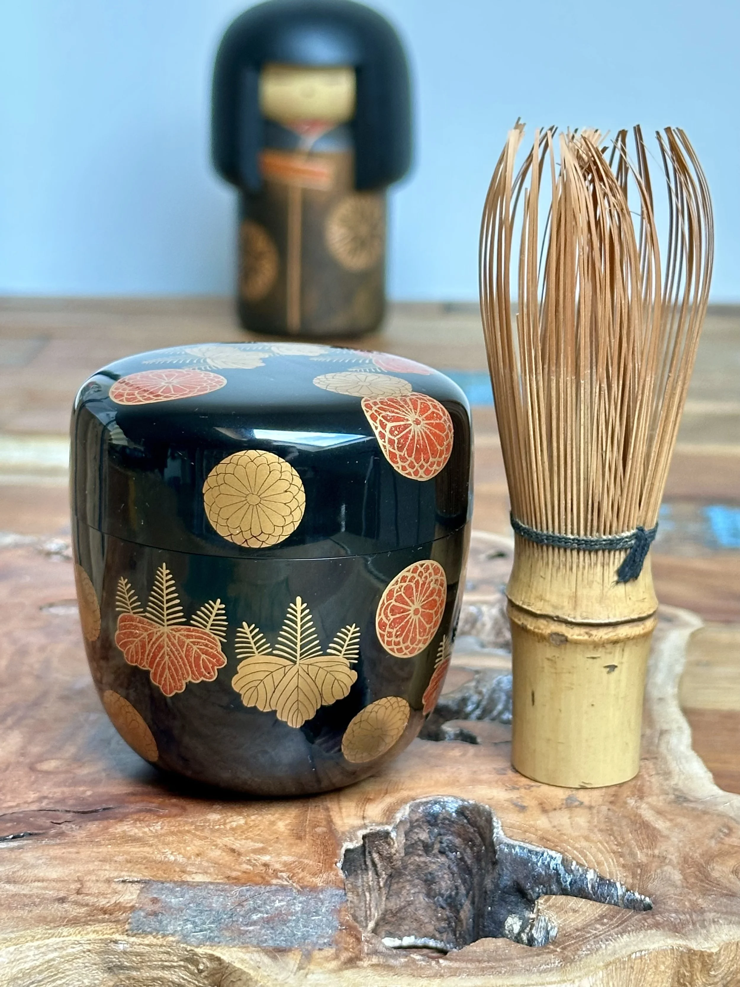 Black lacquered natsume tea caddy with gold and red chrysanthemum and paulownia crests by Yūsai, next to a bamboo matcha whisk on a wooden surface.