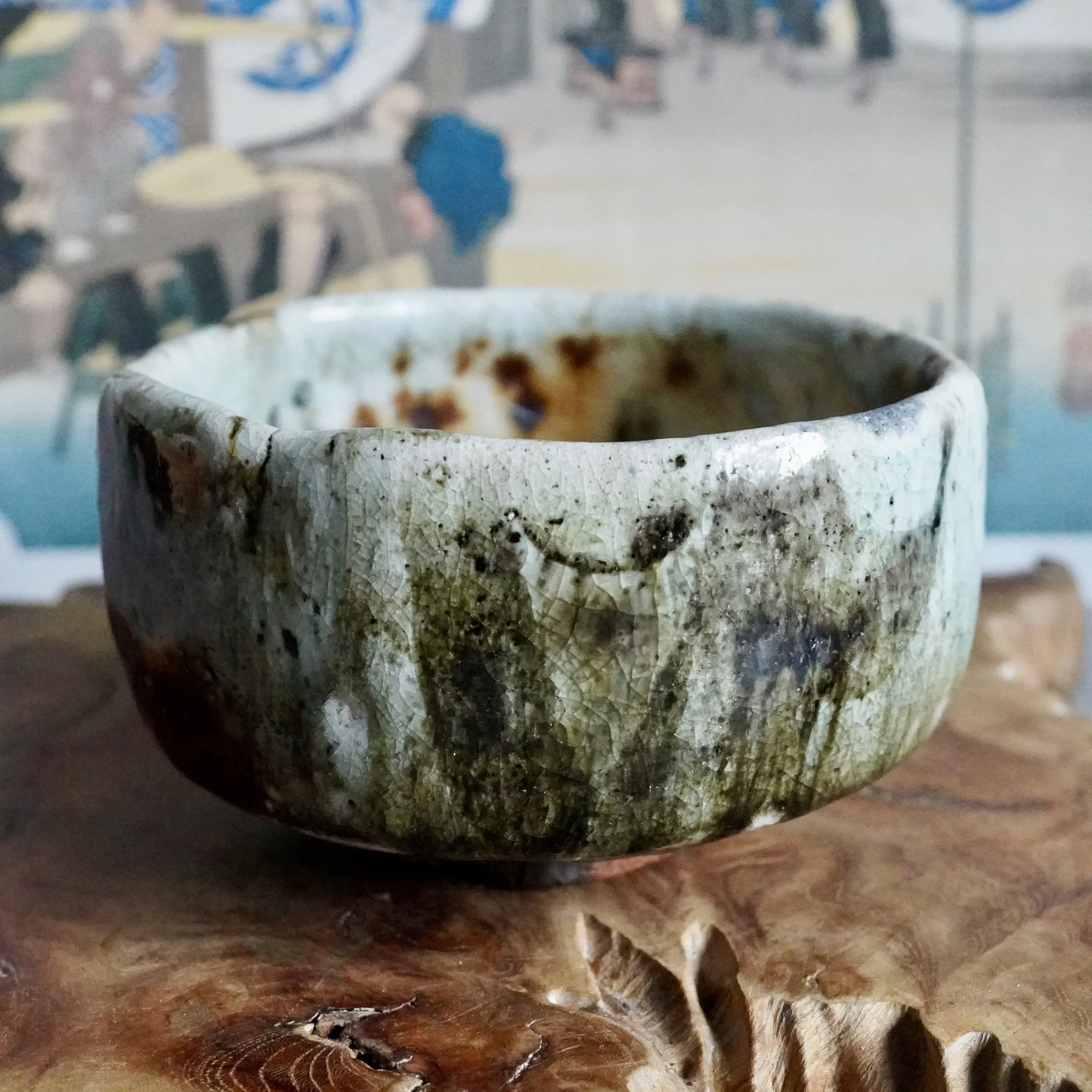 Deguchi Eiji wood-fired chawan with mottled green, rust, and grey crackled glaze on a carved wooden surface.