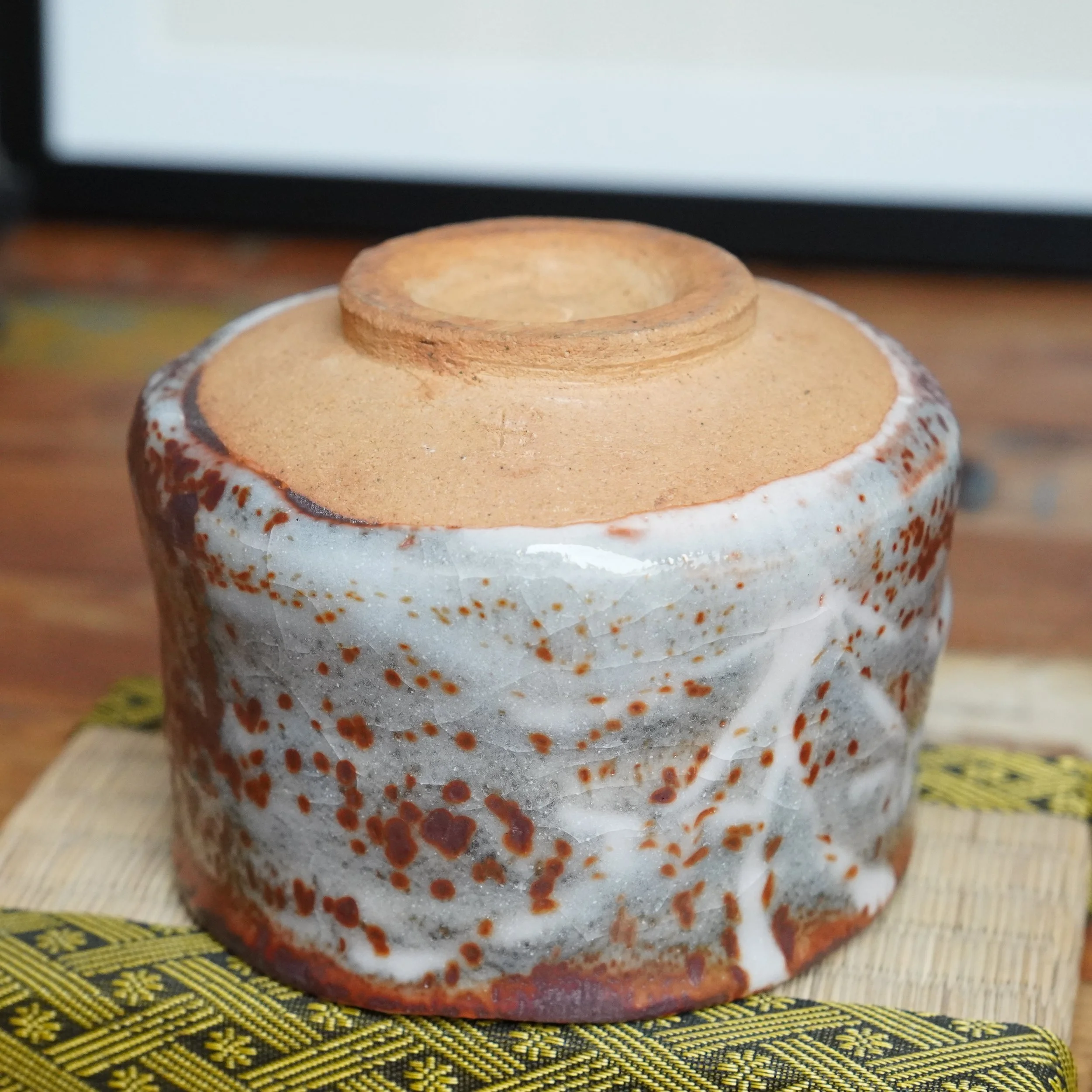 Shino chawan shown upside down, revealing its unglazed light brown foot with an impressed mark. The bowl's body features a crackled white glaze with reddish-brown speckles.