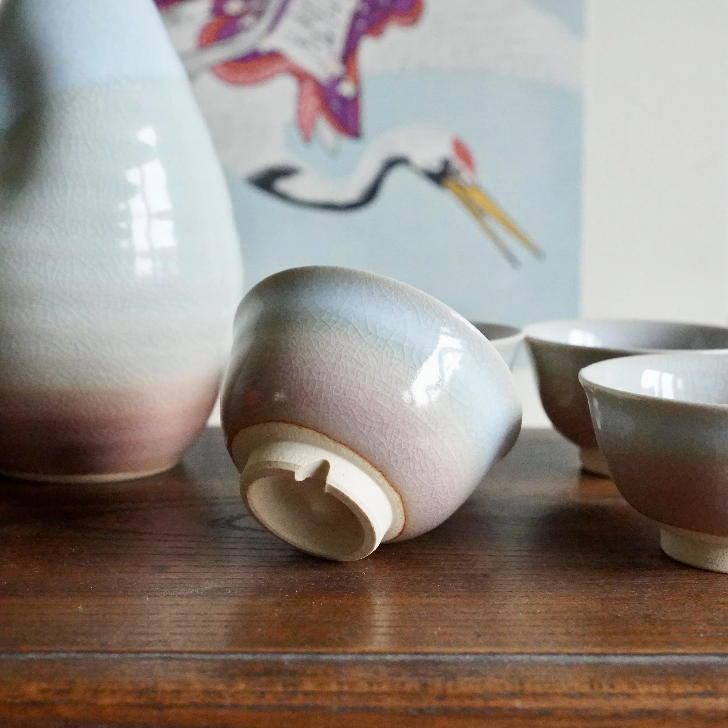 Sake tokkuri and three ochoko cups, with a distinct pale blue-grey to blush gradient and crackled glaze. One cup lies on its side, revealing its notched unglazed foot.