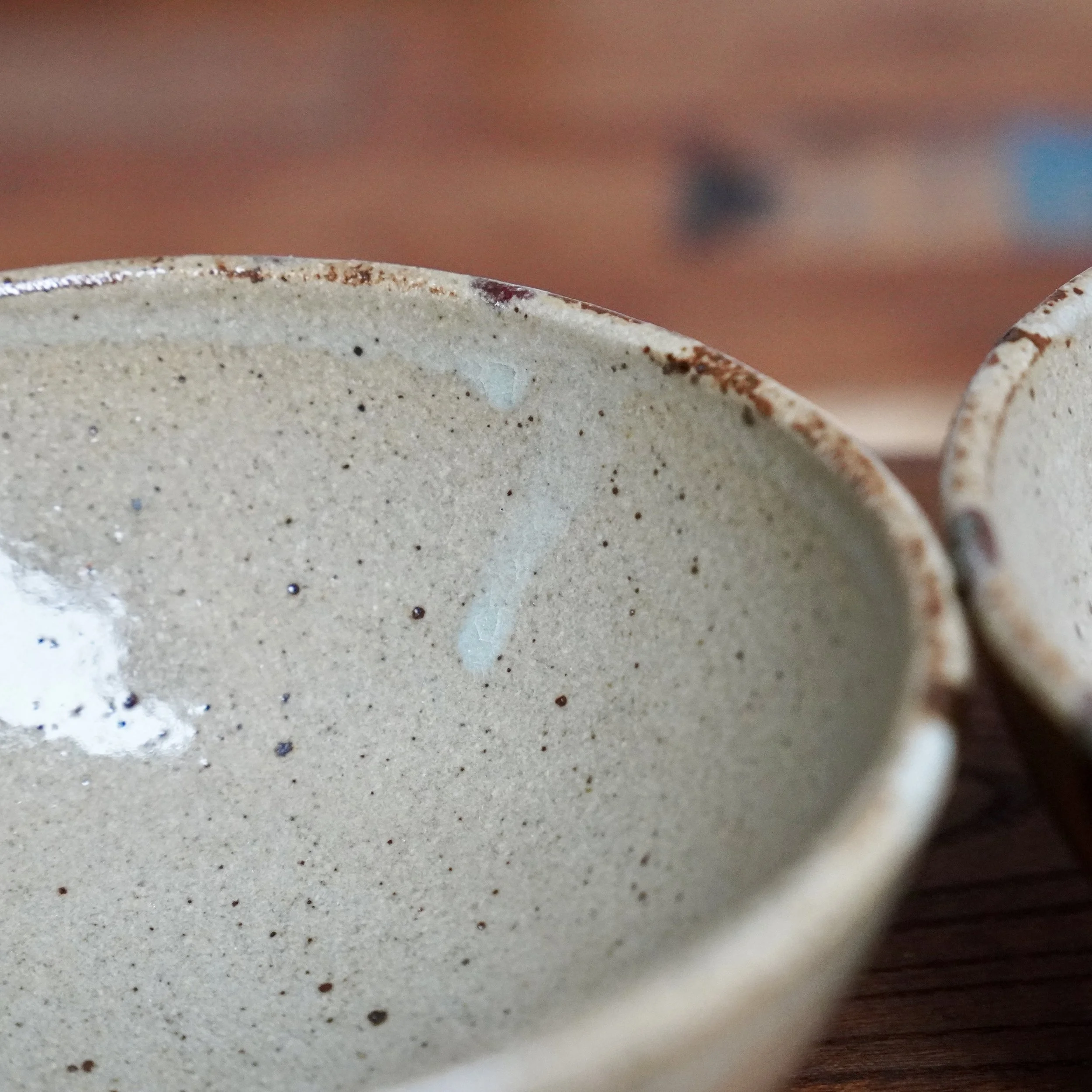 Interior of a pale grey, speckled hira-chawan tea bowl with a reddish-brown rim and subtle milky glaze marks. A second bowl is out of focus to the right.