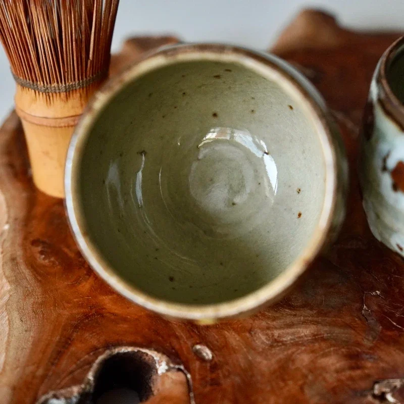 Top-down view into one Kitoen kiln ceramic cup with pale green speckled glaze, a darker rim, beside a bamboo whisk and glimpse of another cup on wood.