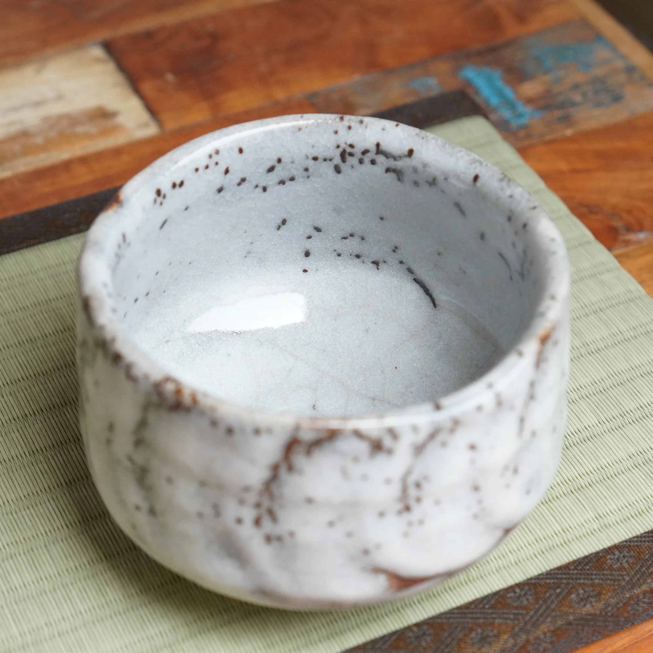 Oni-Shino chawan by Katō Shōzan. White, irregularly shaped ceramic bowl with rust brown speckles on its thick glaze, resting on a ribbed green mat, with a blurred wooden background.