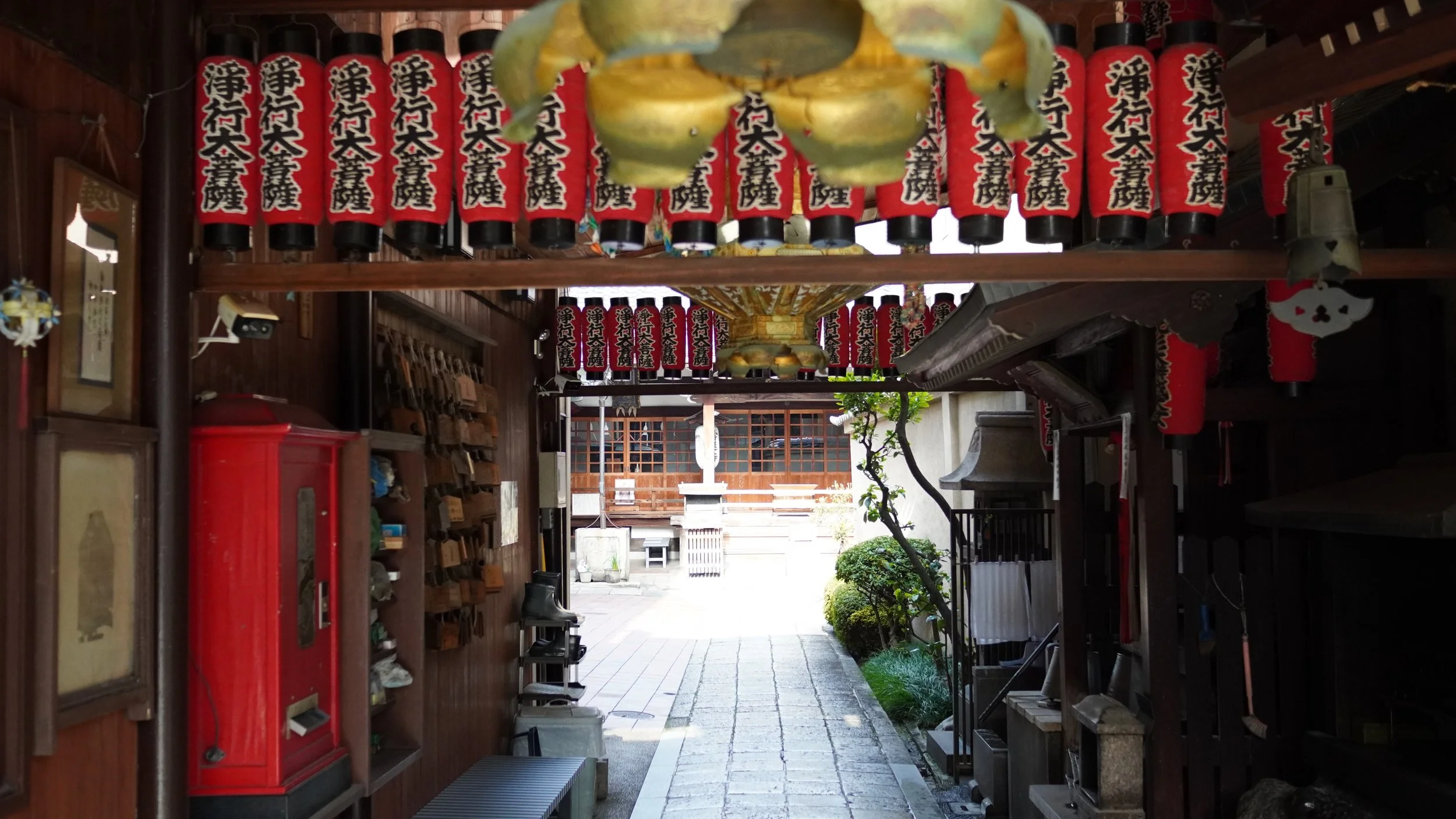 View of the entrance to a traditional Japanese shrine or temple with red and black lanterns hanging overhead. There is a small courtyard area with a tree and stone lanterns in the background.