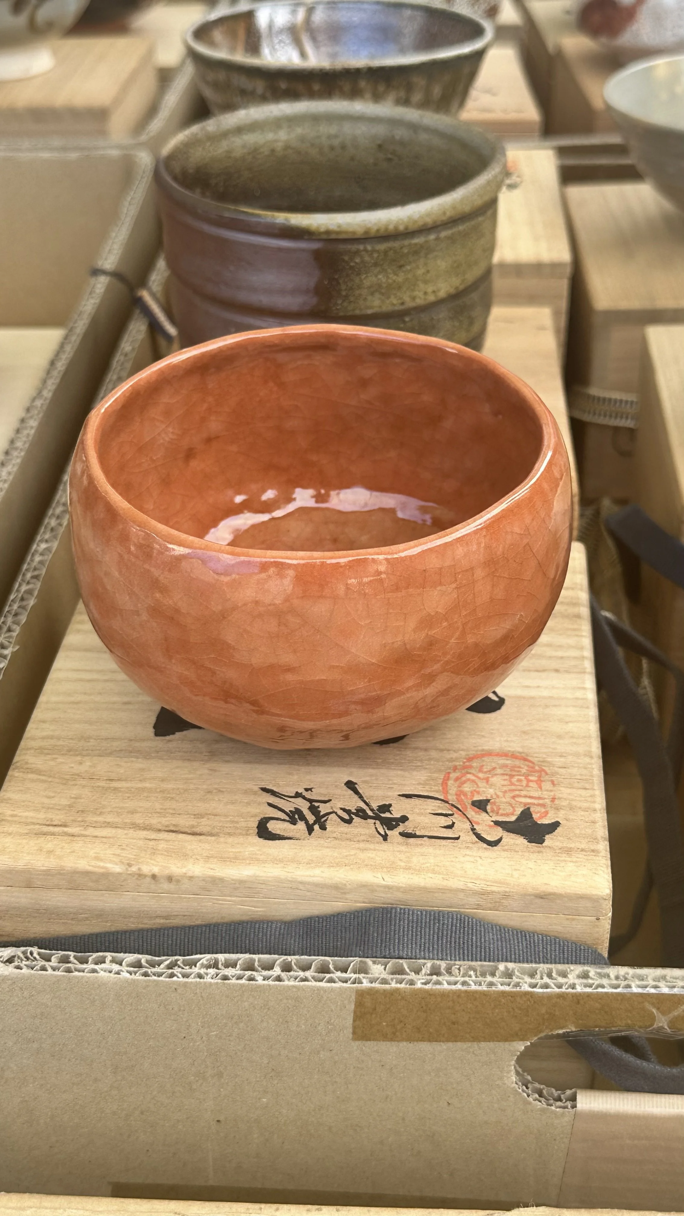 A close-up of a handmade orange chawan matcha mug placed on top of its signed wooden tomobako box in Osaka.
