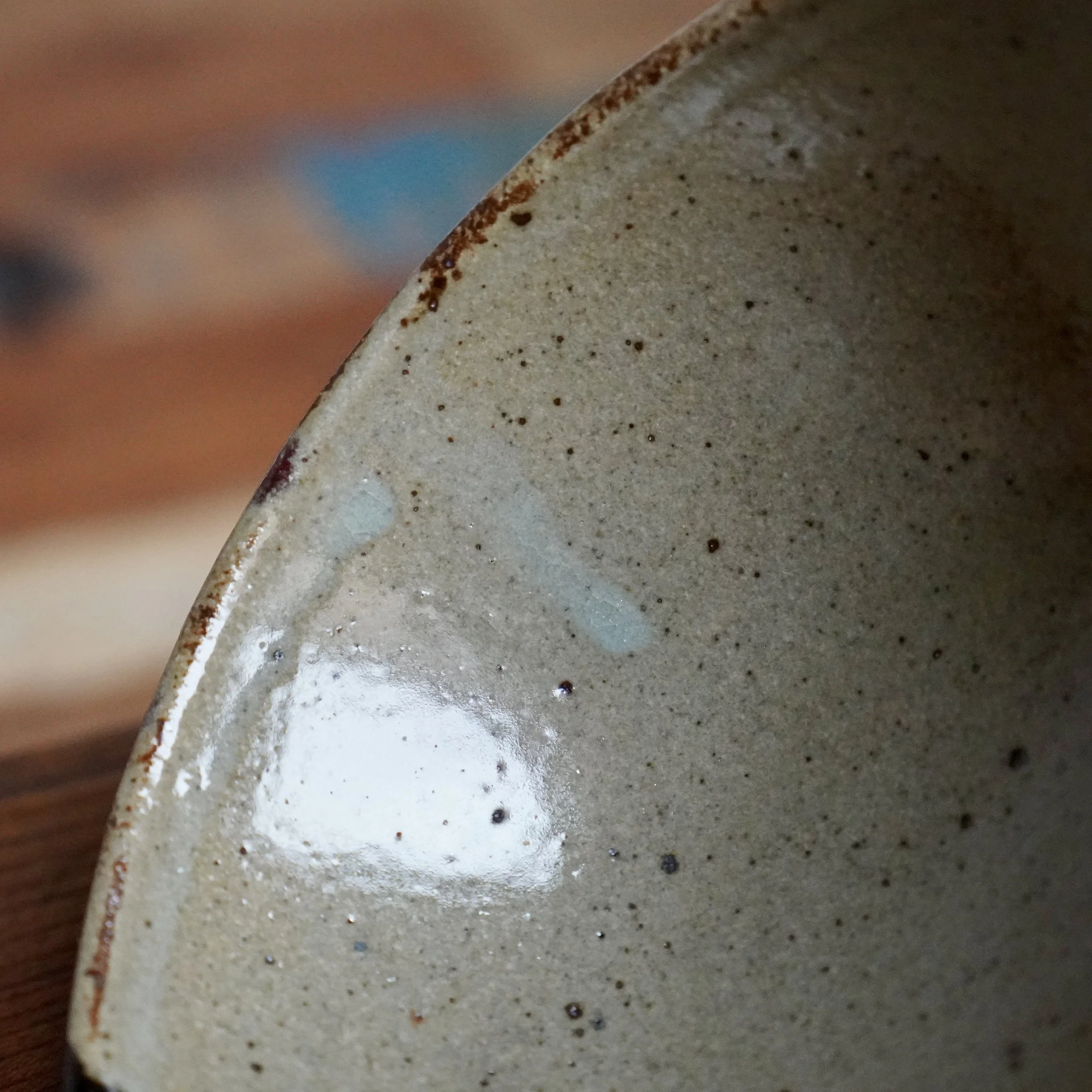 Close-up of a Maruta Nobumasa hira-chawan with a speckled pale grey interior, milky blue-white glaze patches, and a rough brown rim.