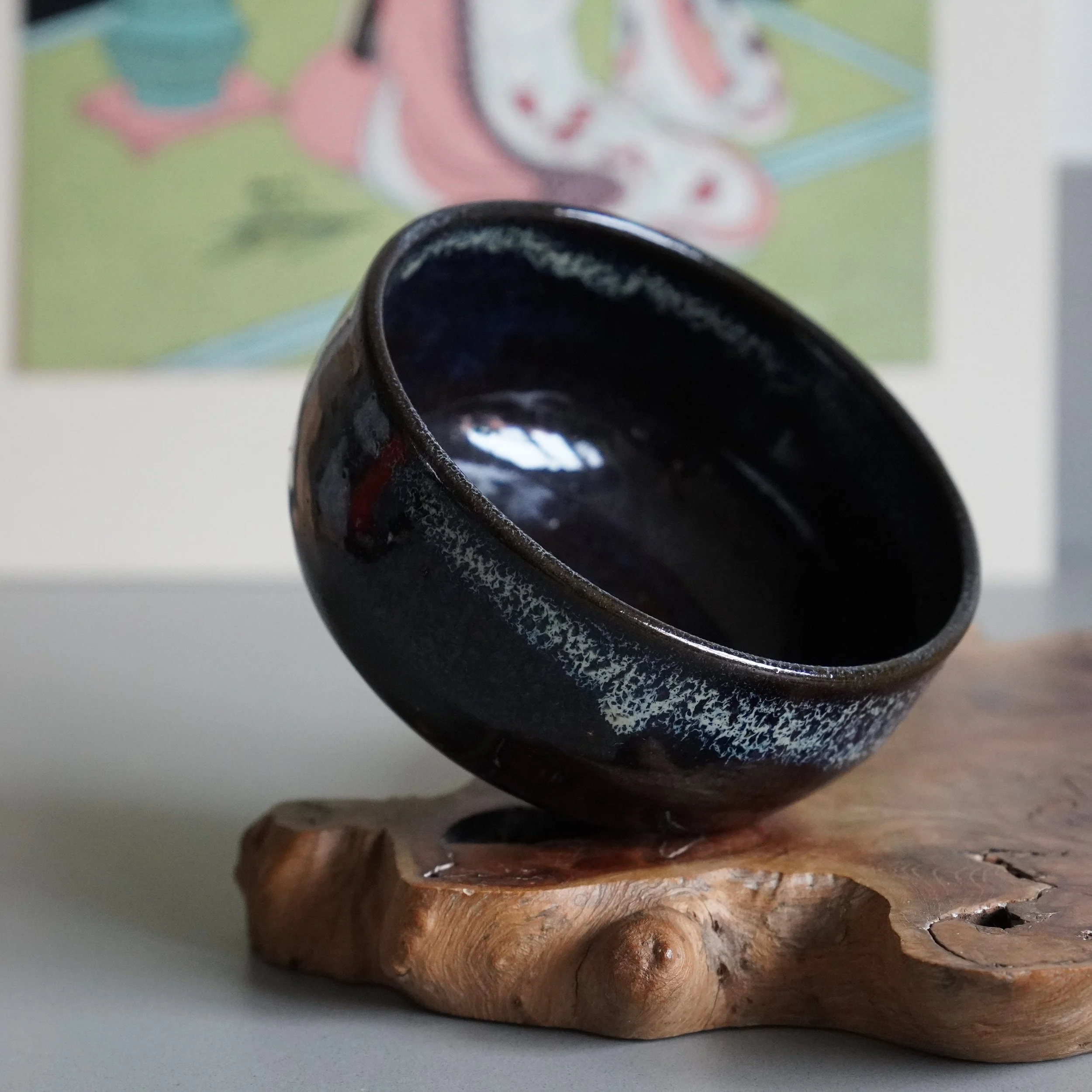 Dark ceramic matcha chawan with textured, blue-black glaze and white speckles, tilted on a natural wood stand.