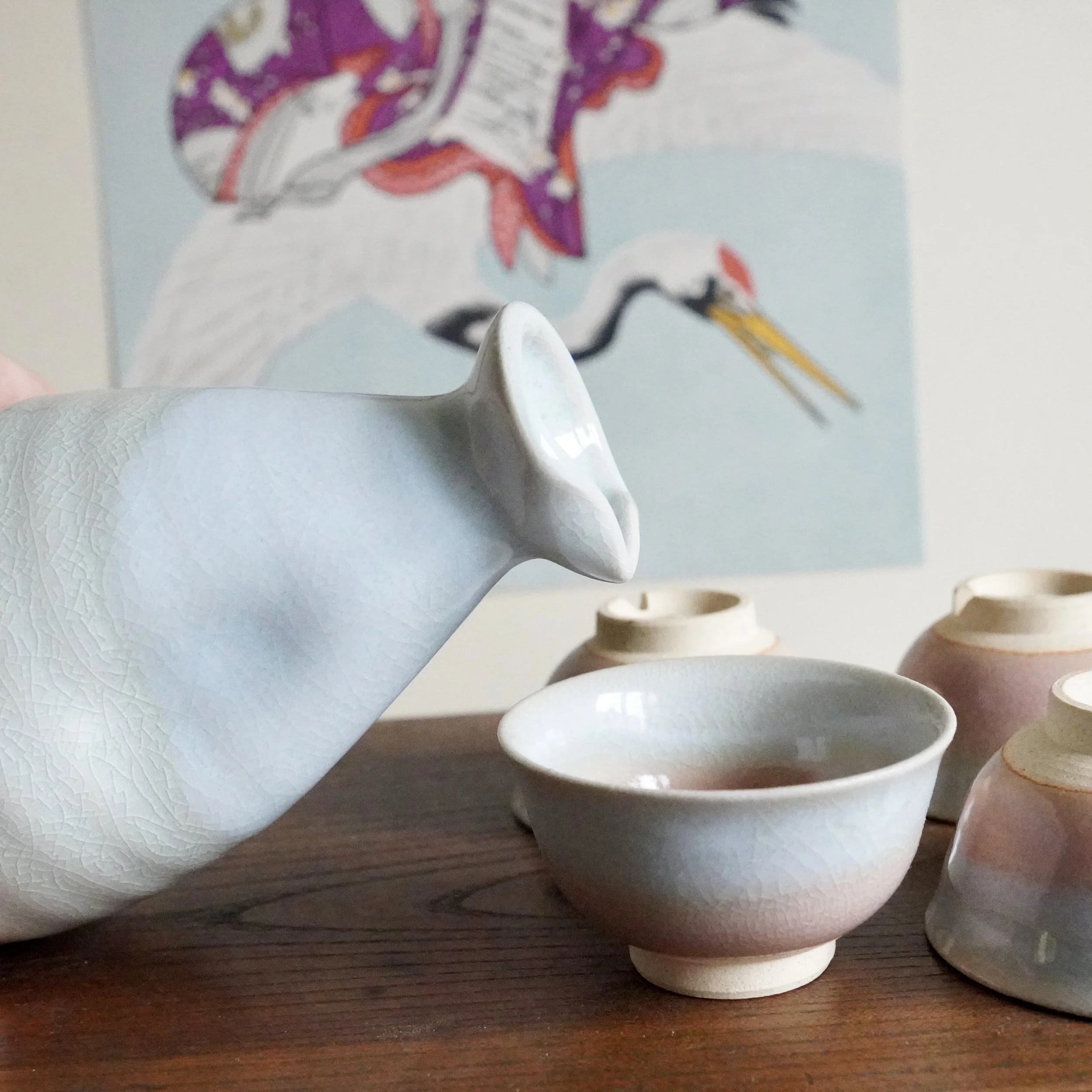 Hagi-yaki tokkuri pouring liquid into an ochoko, both with crackled blue-grey and pink gradient glaze, on a wooden surface with blurry art behind.