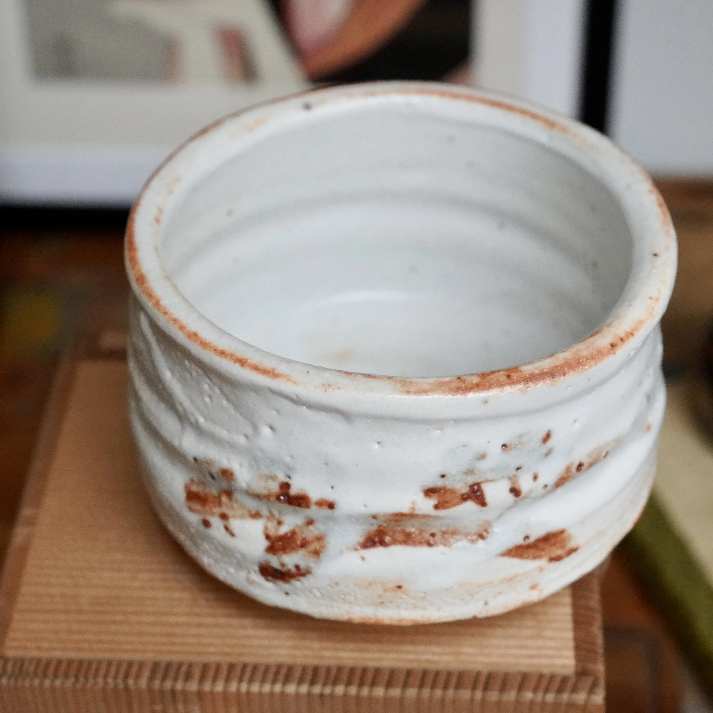 Signed Shino chawan with irregular white glaze, horizontal ridges, and rust-orange marks on the rim and body, resting on wood.