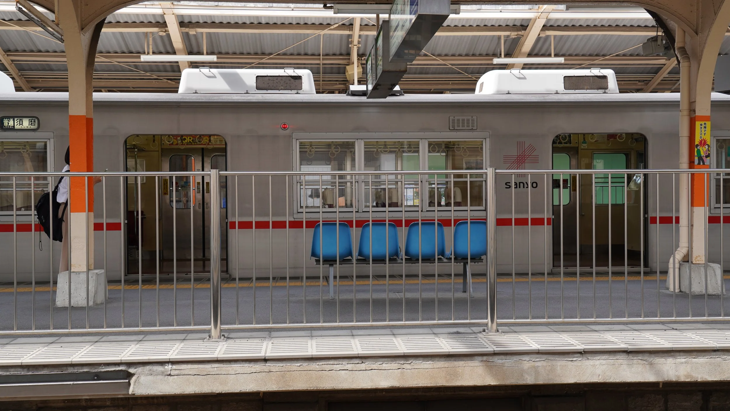 A silver Sanyo Electric Railway train stopped at a quiet Japanese train station platform with blue waiting seats and orange pillars during a sourcing trip.