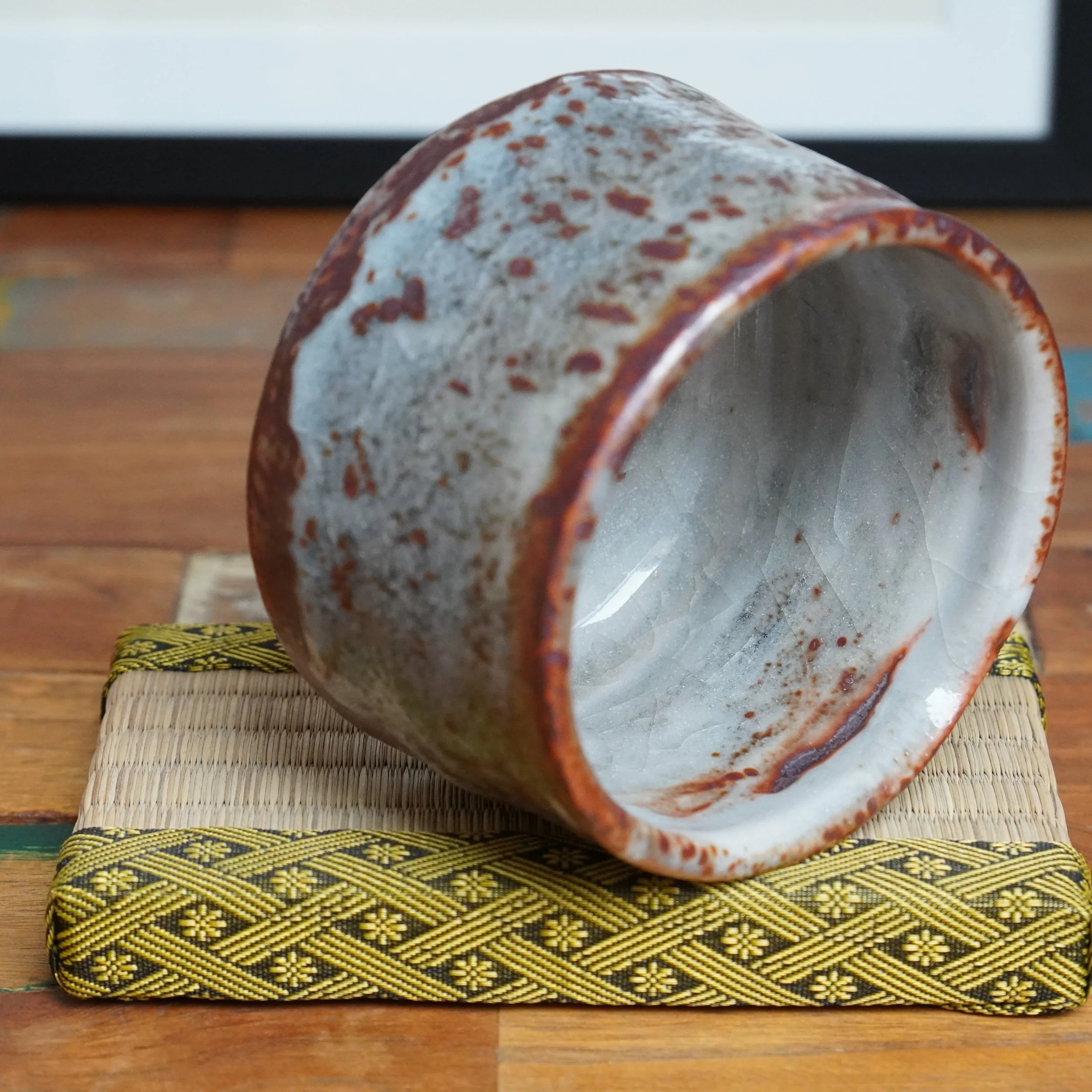 Shino chawan lying on its side, showing pale grey glaze with reddish-brown speckles and crazing inside. It rests on a straw mat with a gold and black patterned border.