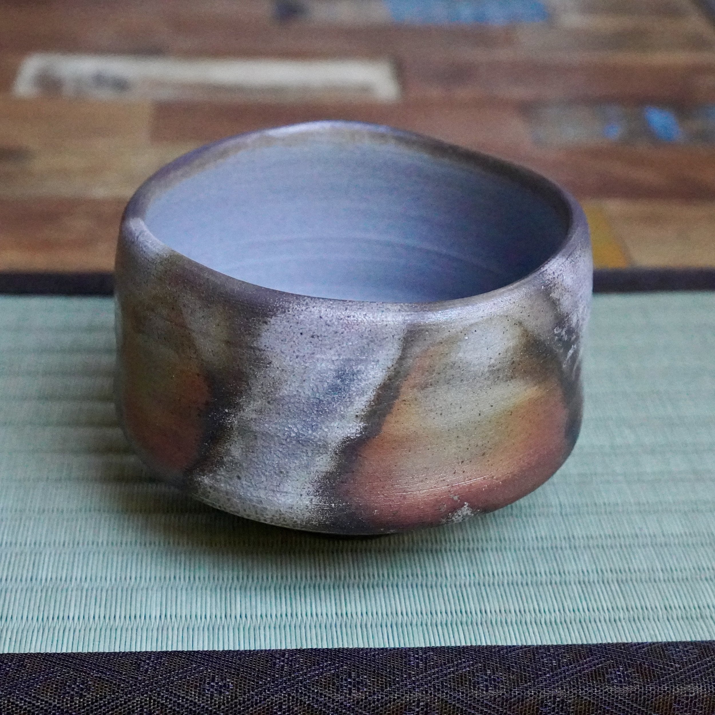 Inomata Masaaki's Sangiri Bizen chawan with unglazed ash grey, rust, and burnt orange exterior, and light blue-grey interior, on a green tatami mat.