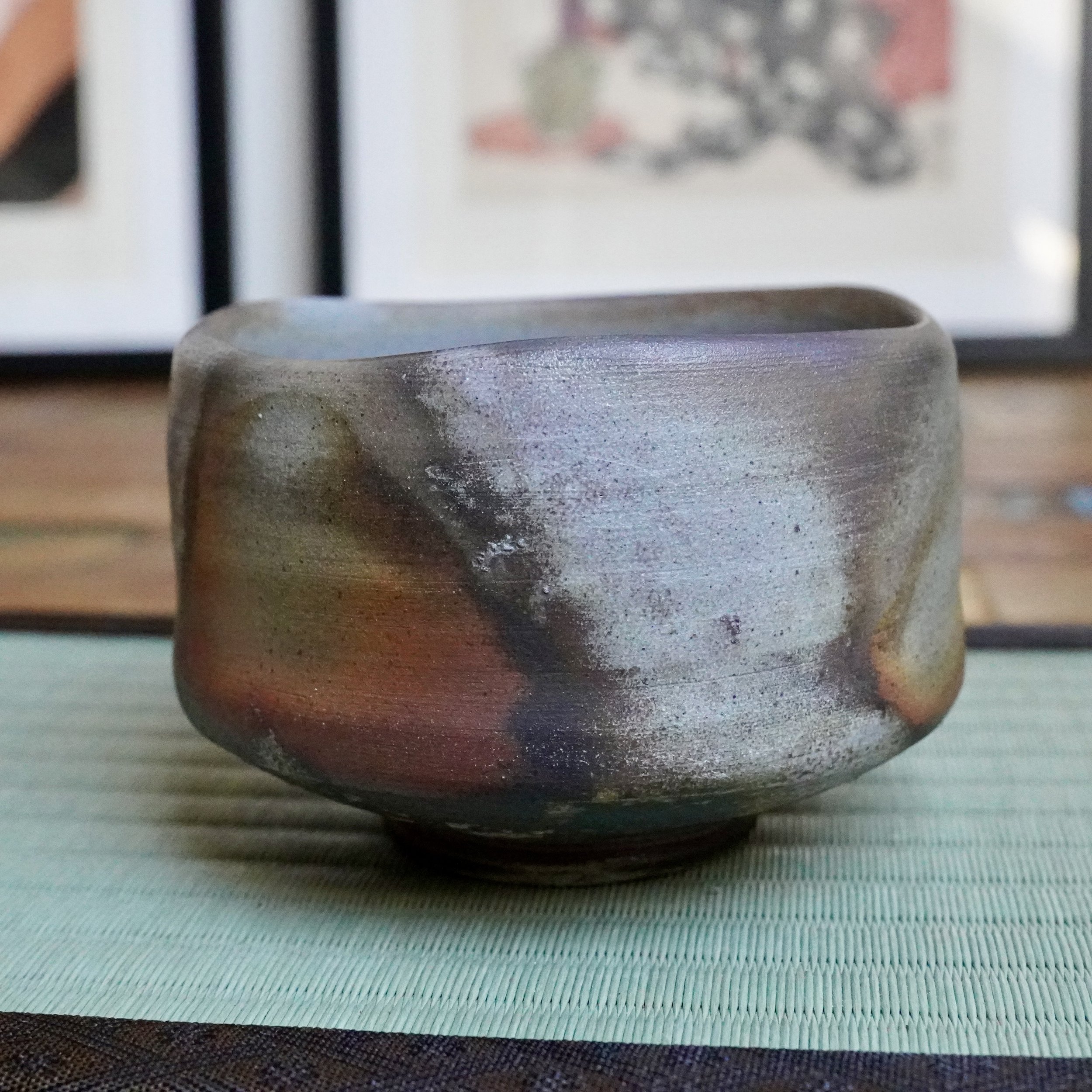 Inomata Masaaki's Bizen chawan, showing rust and burnt orange bleeding into ash grey and silvery tones, resting on a green woven mat.