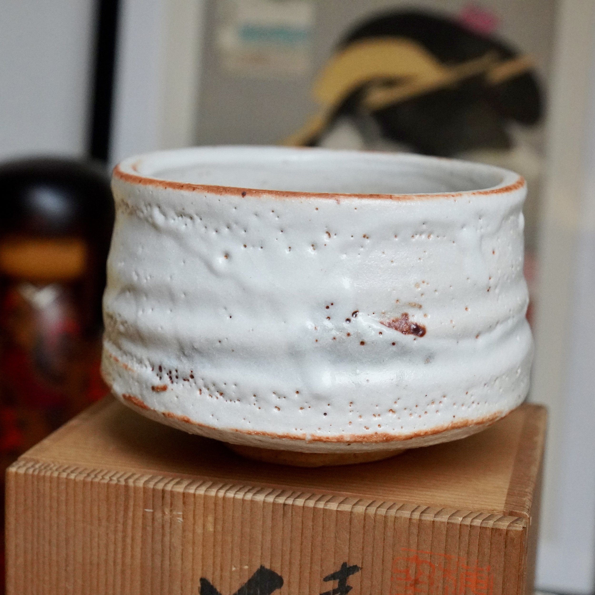 Textured white Shino chawan with reddish-orange rim and surface marks, resting on a light wood tomobako with joinery and dark script.