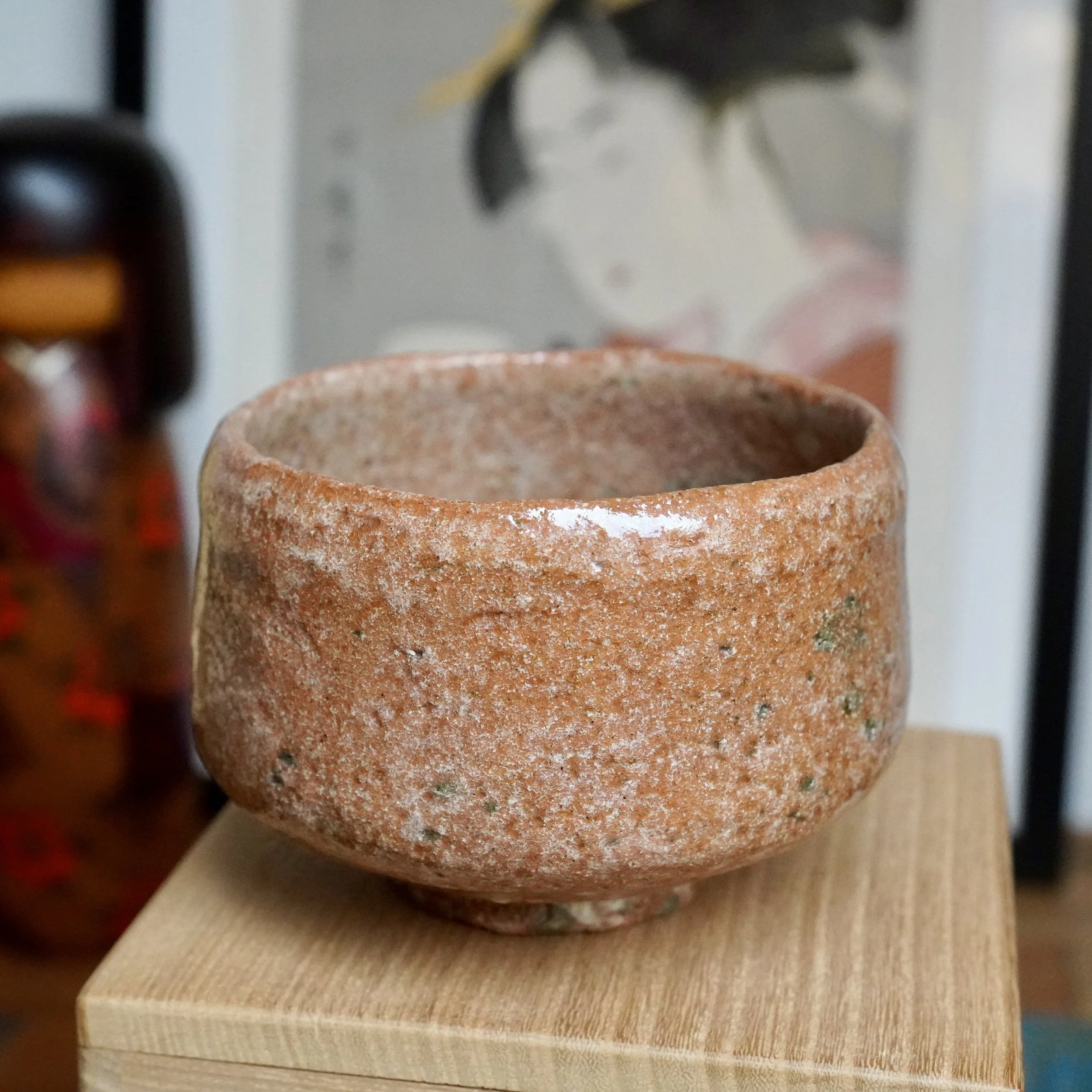 Sawada Hiroyuki's aka-raku chawan, reddish-brown with a rugged, pitted surface, white glaze drip, and green specks, resting on a wooden box.