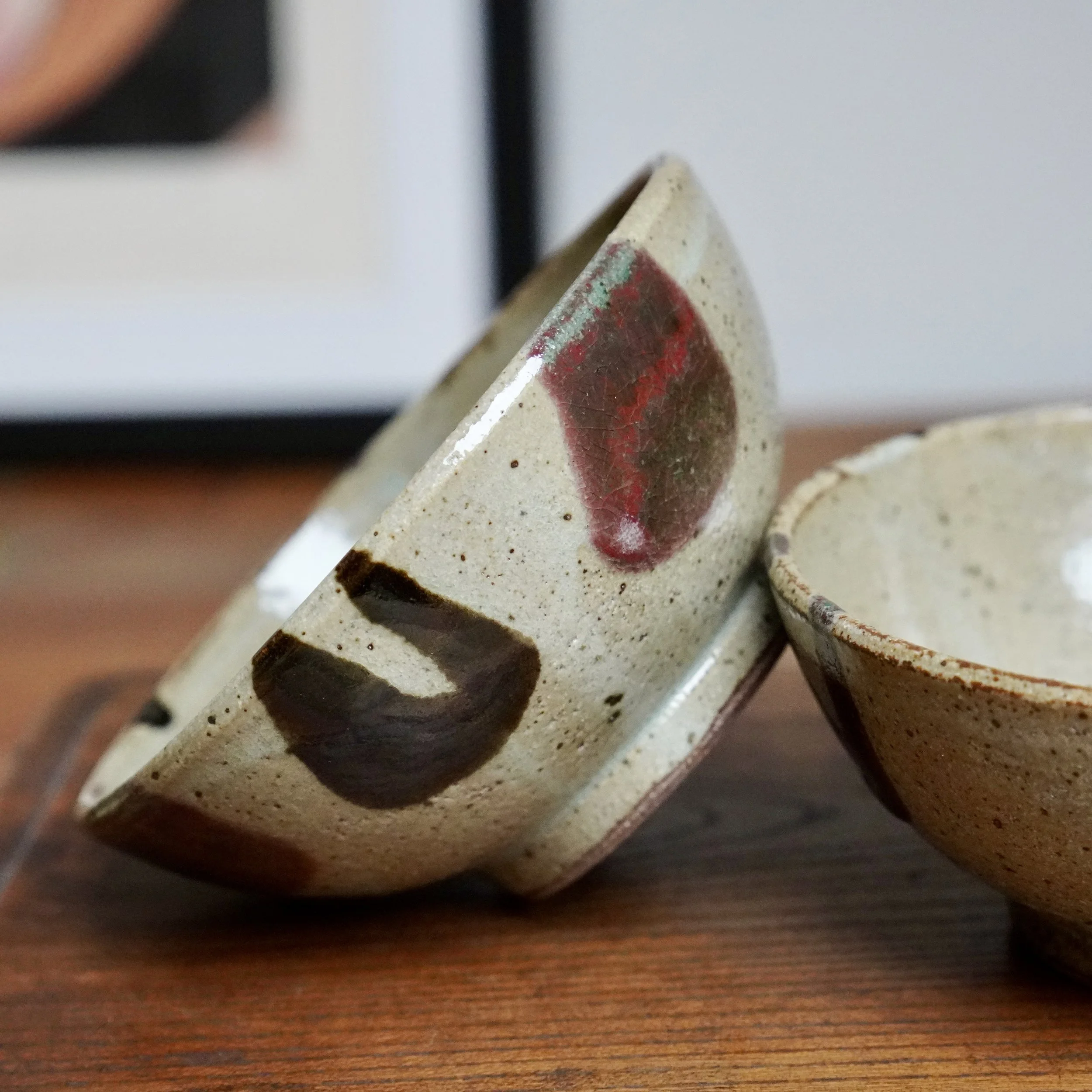 Pair of speckled tea bowls with milky glaze drips, dark brown brush marks, and crackled red patches, resting on a wood surface.