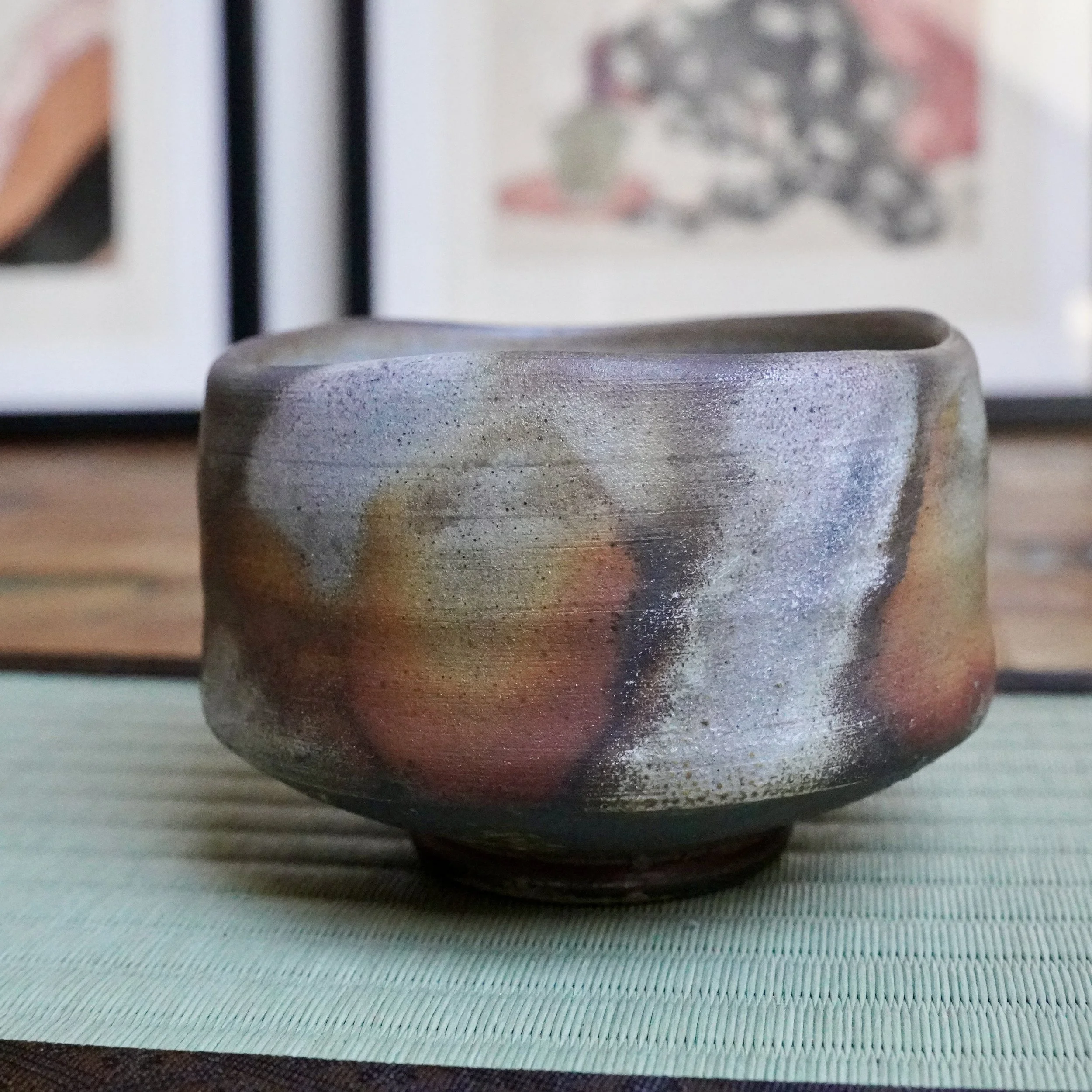 Inomata Masaaki's Bizen chawan, irregularly shaped, showcasing sangiri effects of rust and orange fading into ash grey and silver, on a green woven mat.