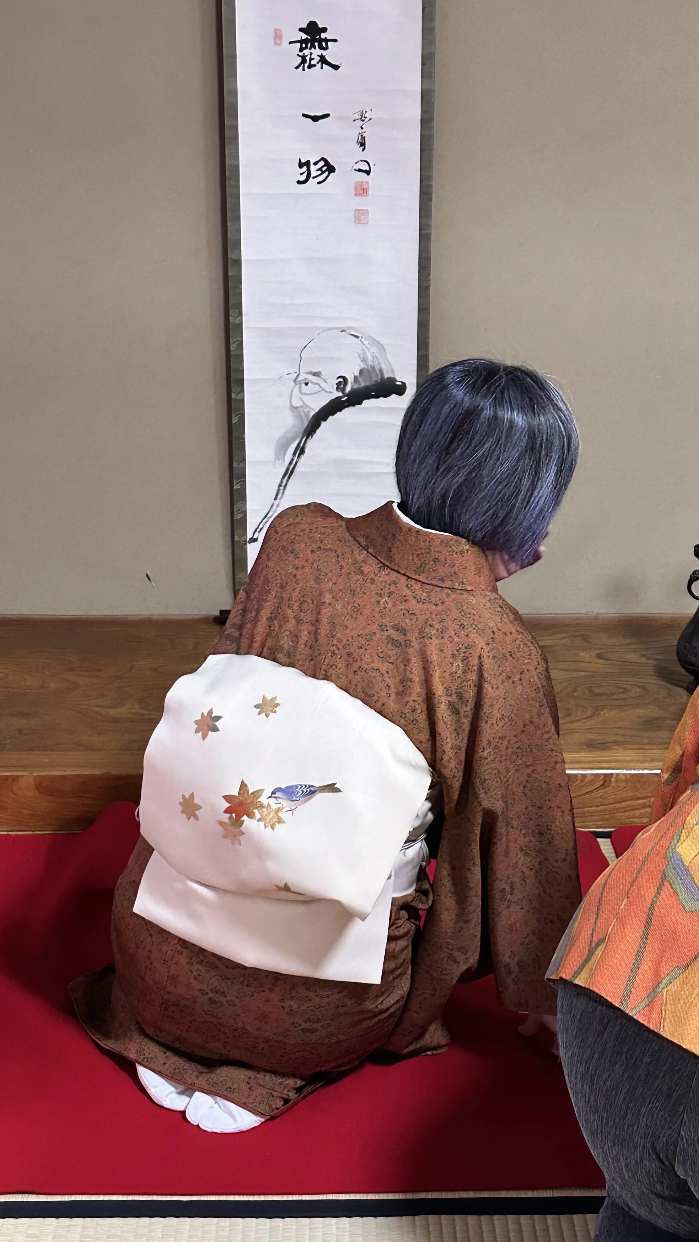 Woman in brown kimono seated in seiza before a Japanese kakejiku hanging scroll with Zen calligraphy reading mu ichi motsu, "not one thing", and an ink painting of a Zen monk. Autumn obi with maple leaves and bird motif. Japanese tea ceremony.