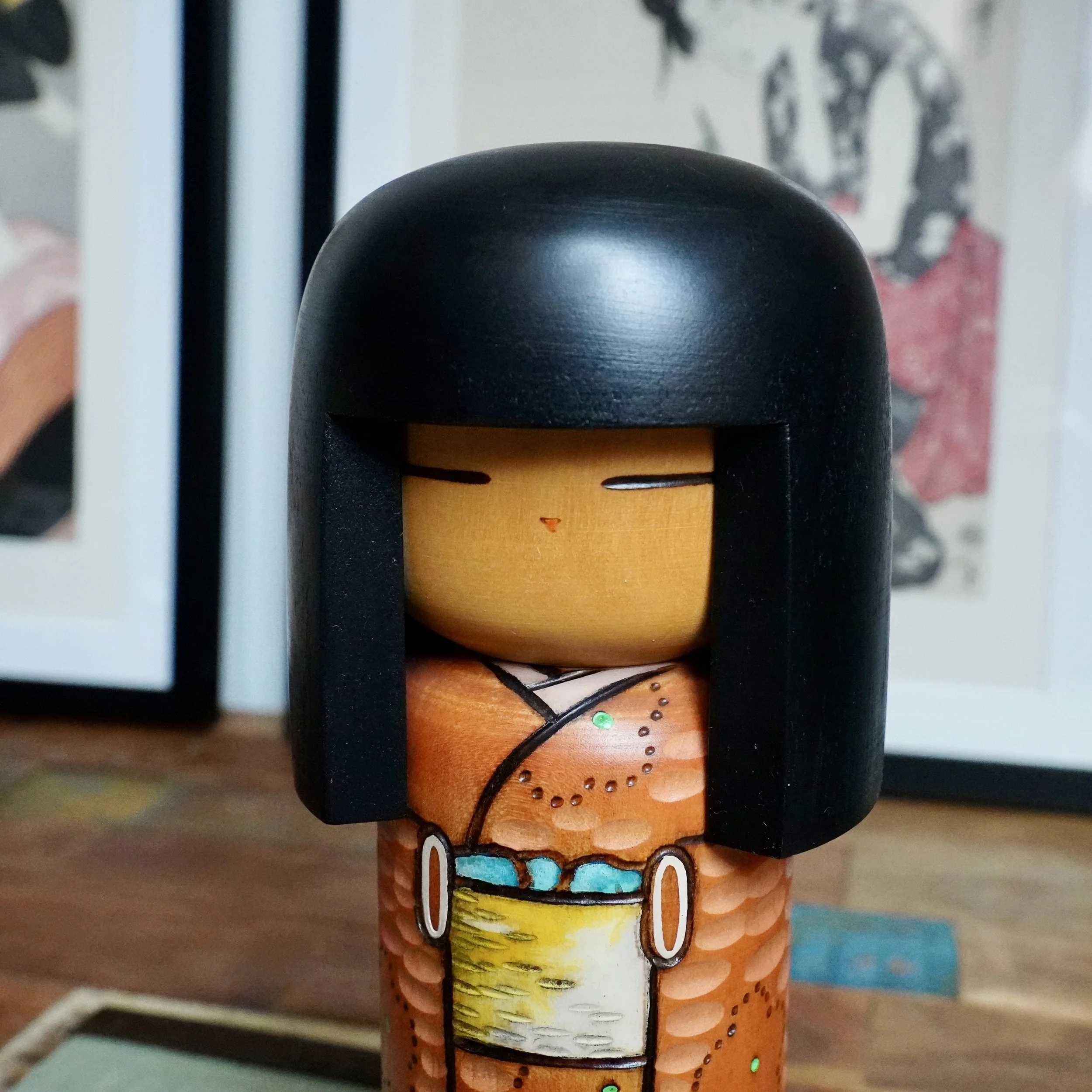 Kisaku sōsaku kokeshi doll, front view, with black hair, simple painted face, and carved orange-brown kimono decorated with ume blossoms and a pale yellow obi banded in turquoise.