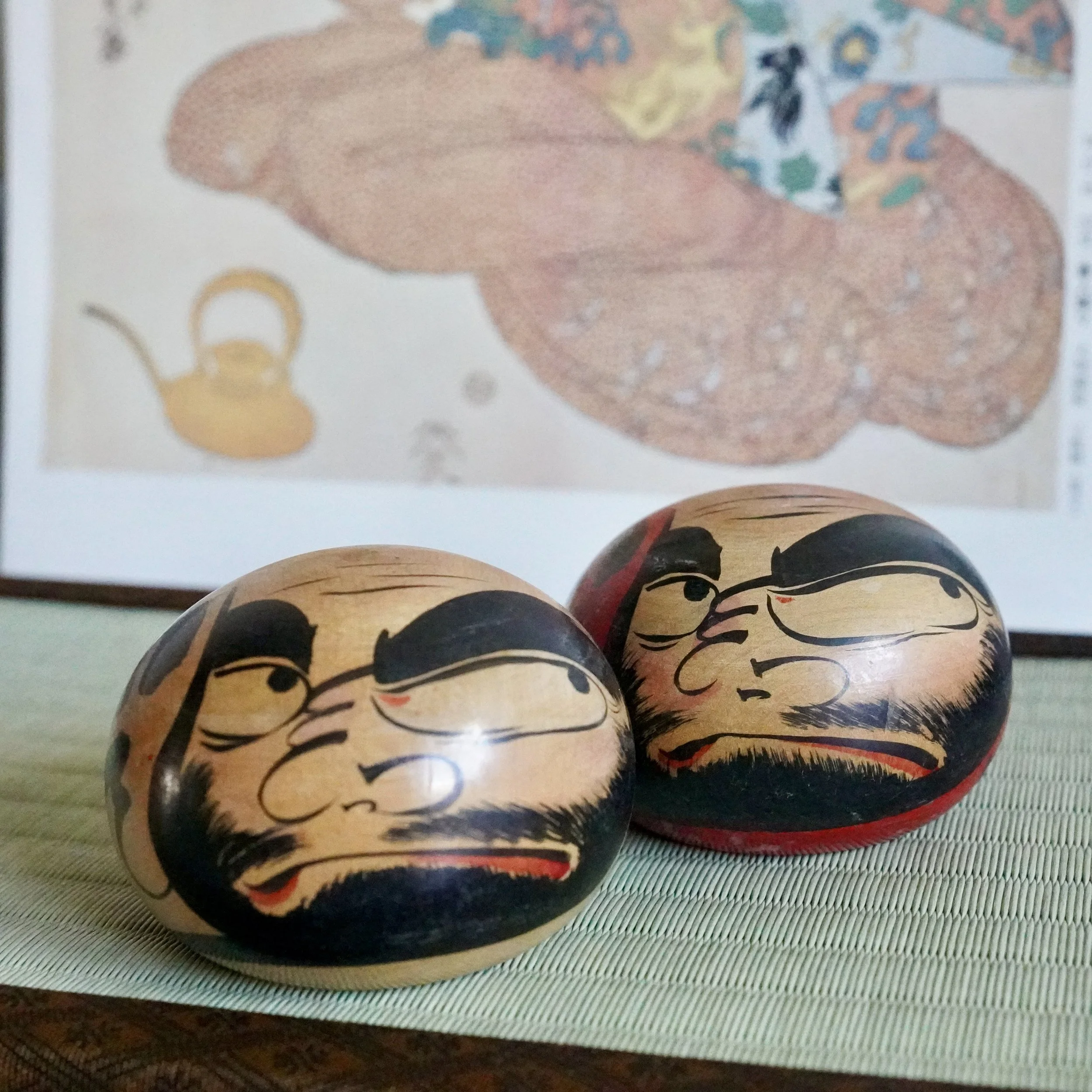 Satō Zenji's Tsugaru daruma pair: two wooden dolls with hand-painted fierce eyebrows and downturned mouths, one with a red base, on a tatami mat.