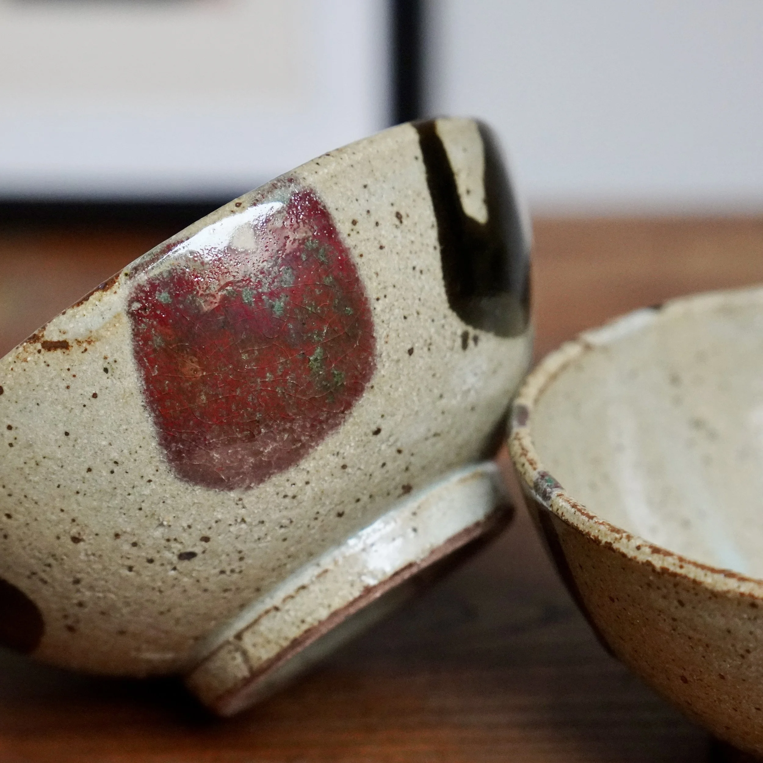 Two hira-chawan from Maruta Nobumasa kiln. The front bowl is angled, showing a freckled pale grey exterior with a crackled wine-red glaze and a black brushstroke. A second blurred bowl is visible.
