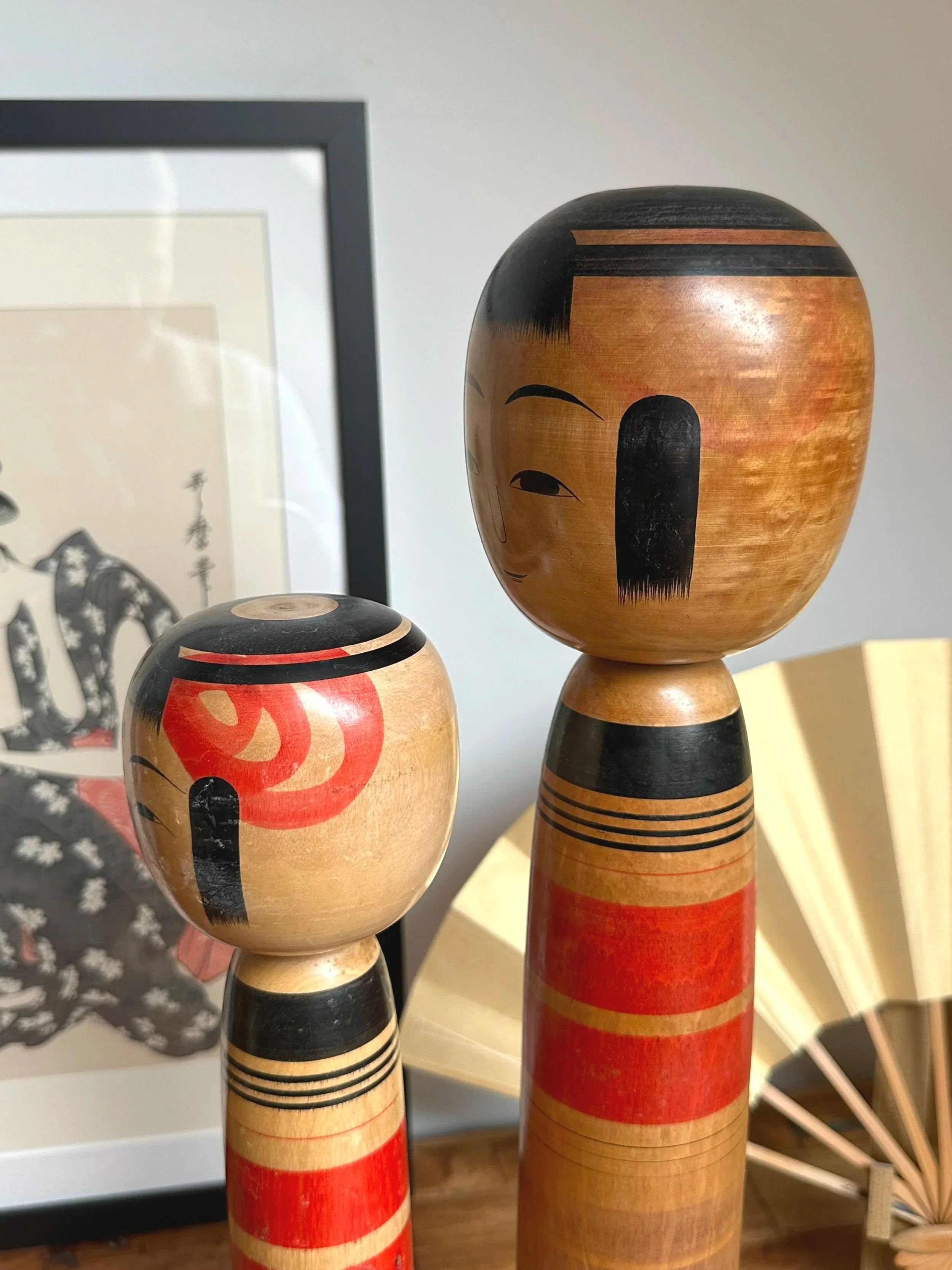 Two Tsuchiyu Kokeshi dolls, one taller, with red and black bands on natural wood bodies. The smaller doll's head features a red and black swirl.