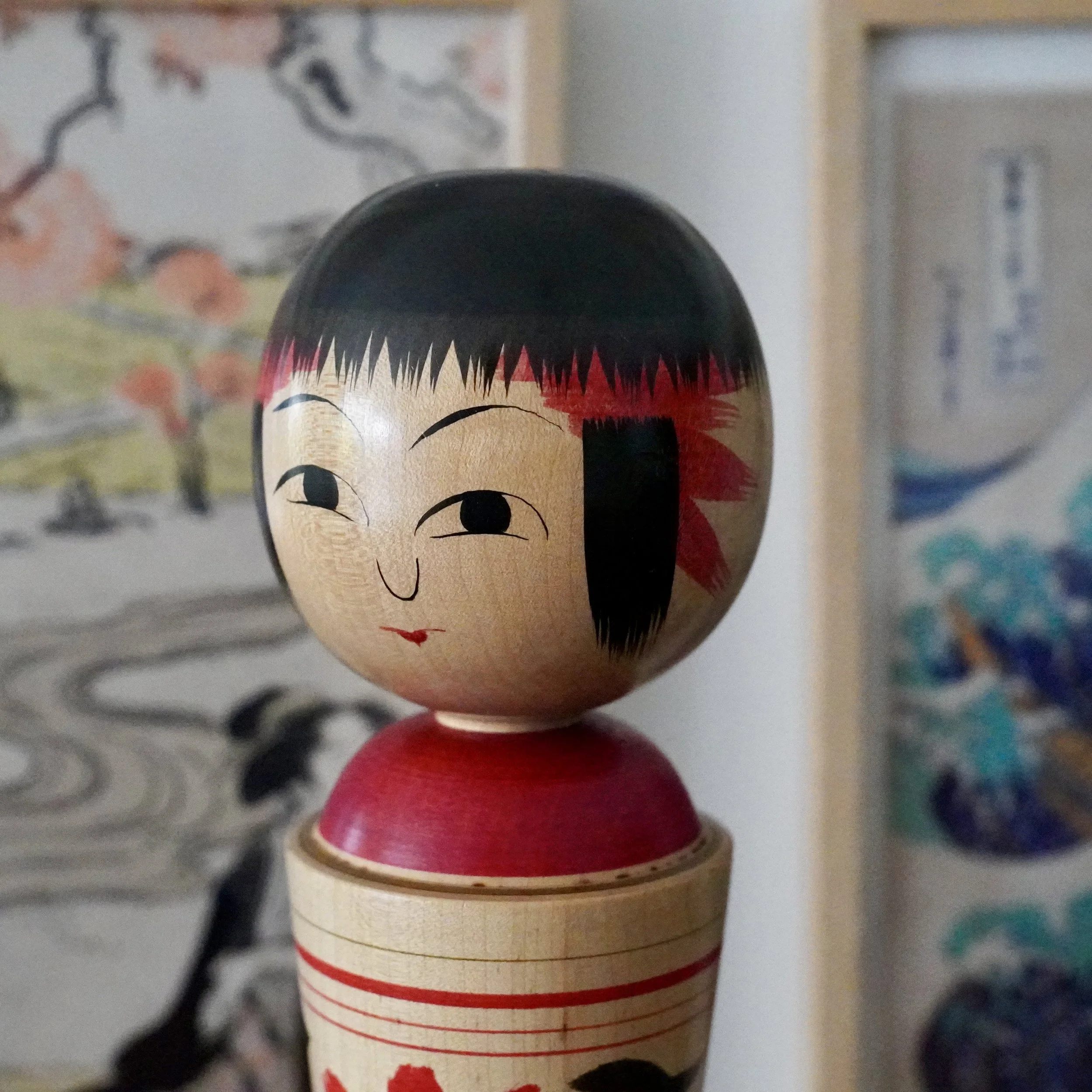 Close-up front view of a Kokeshi doll by Izu Mamoru. Light wooden face, black bob hair with red spiky design, painted eyes, nose, and mouth. Red collar and striped body base.