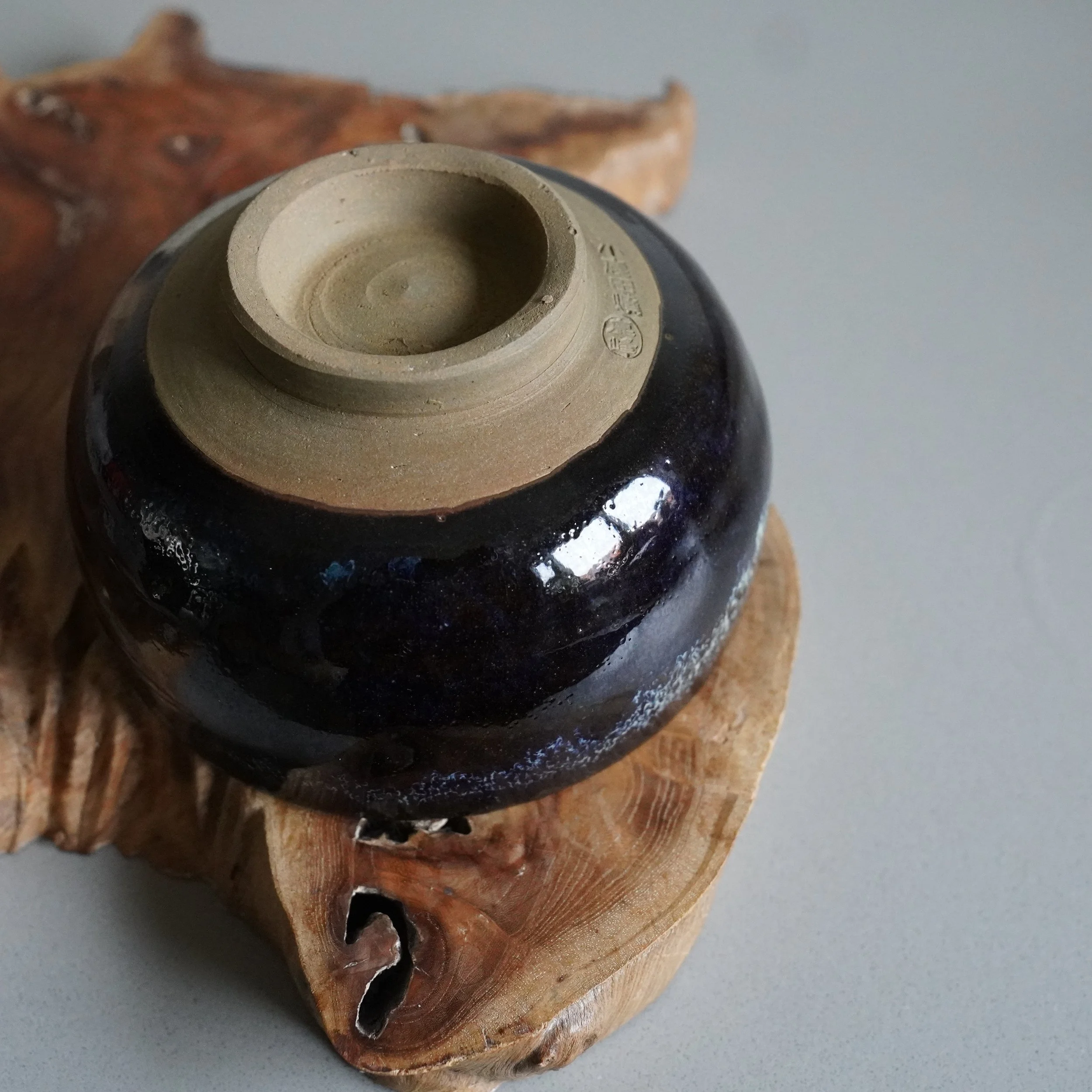 Underside of an Onta-yaki matcha chawan, dark blue-brown glazed, resting on a carved wooden base. The unglazed foot shows a signature by Rokuhō.