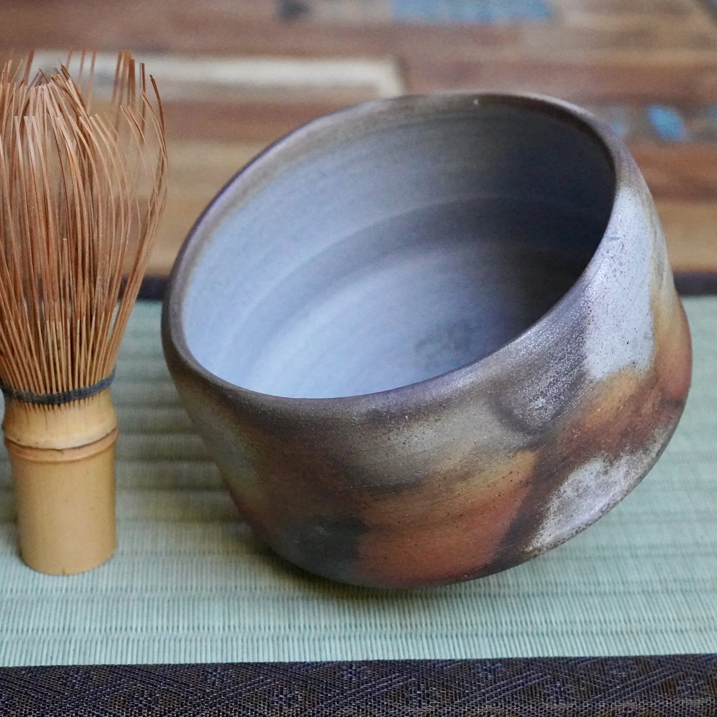 Inomata Masaaki's Sangiri Bizen chawan, featuring a rust and ash-toned exterior and pale blue-grey interior, beside a bamboo whisk on a woven mat.
