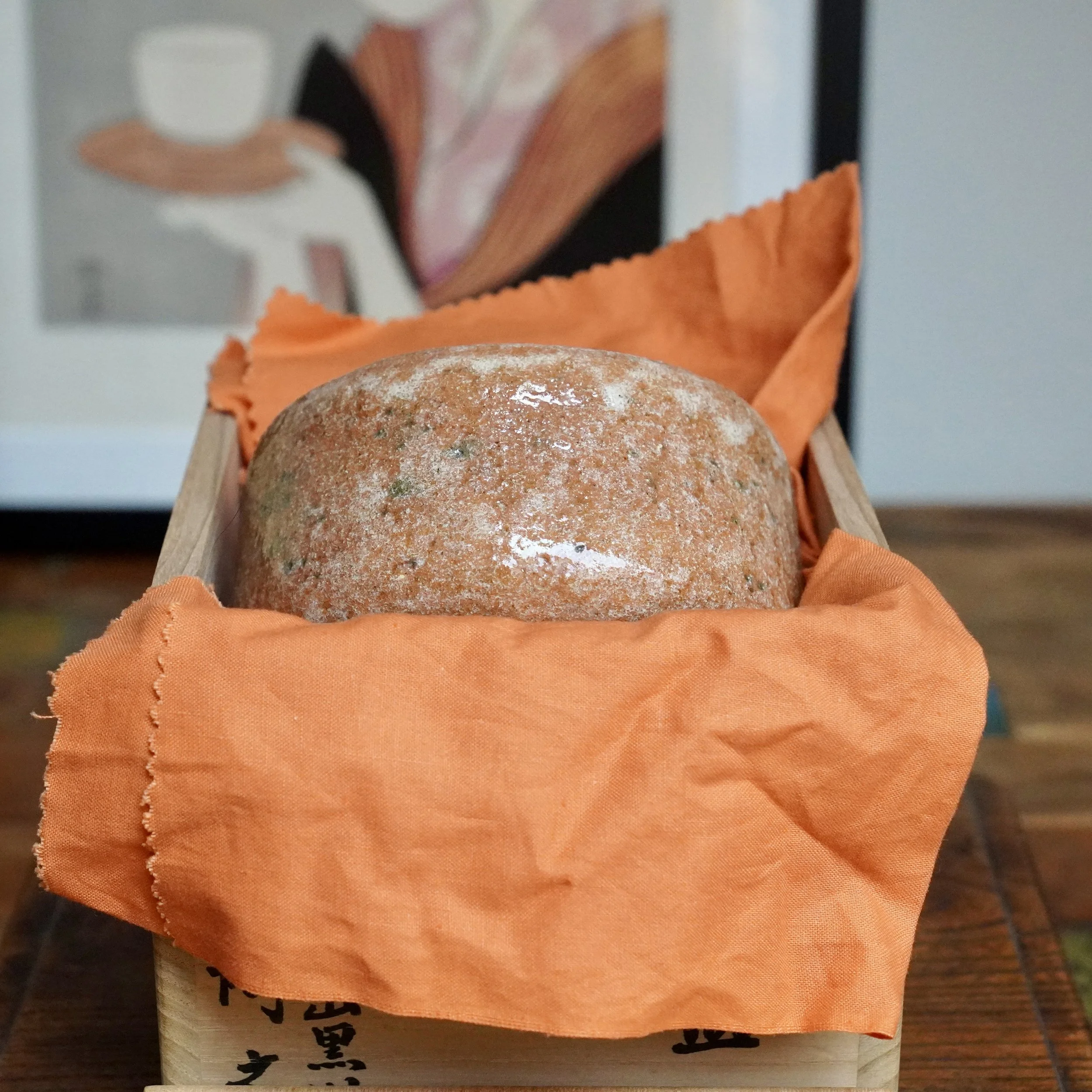 Sawada Hiroyuki's aka-raku chawan, a rugged reddish-brown bowl with glossy patches and a pale glaze drip, nestled in orange fabric in a wooden box with visible kanji.