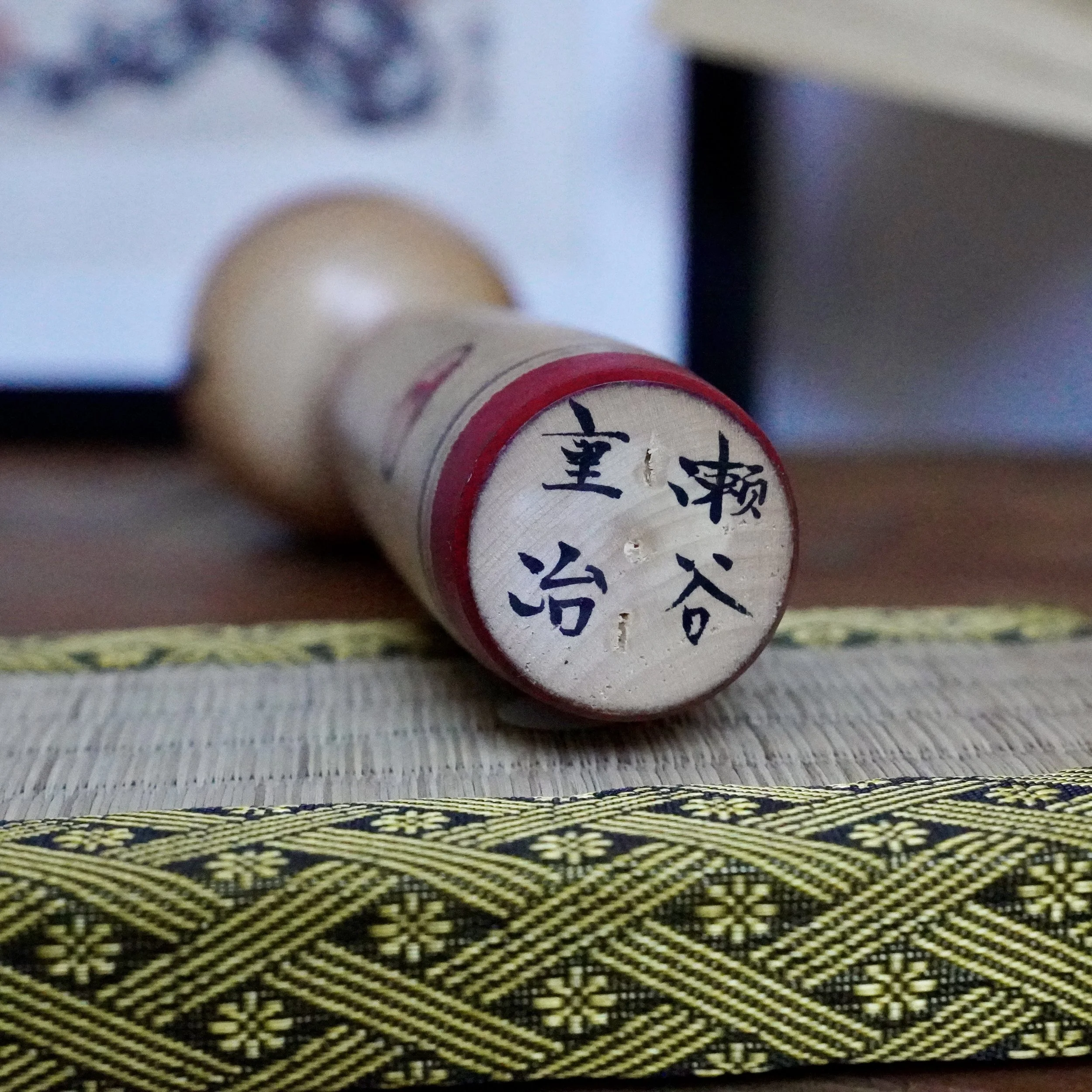Close-up of a wooden Nakanosawa Takobozu kokeshi doll's base, signed "Seya Jūji" in black ink with a red painted band, resting on a woven mat.