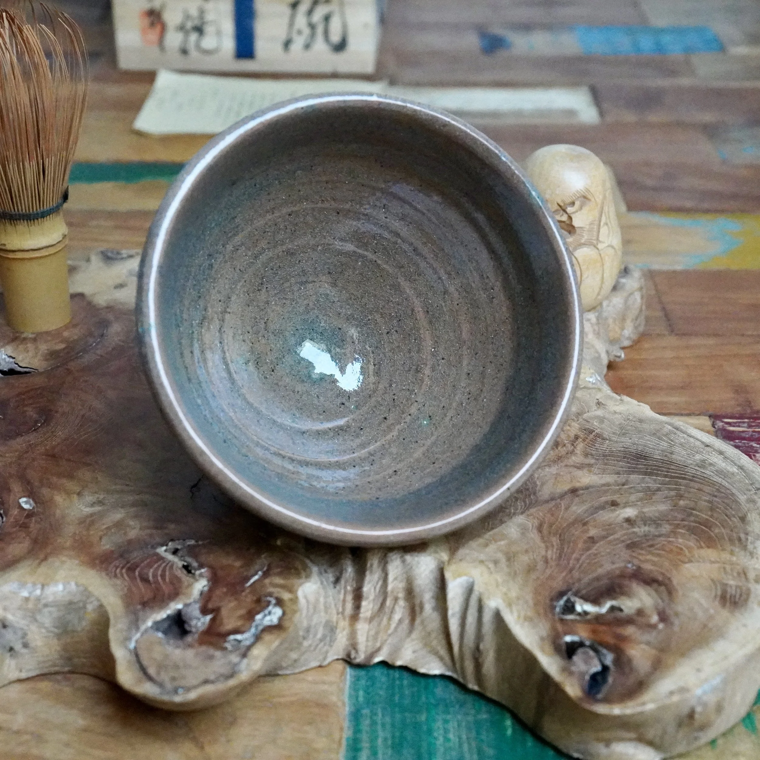 Mizushima Jirō's Kagurao-yaki chawan displays a blue-green speckled glaze inside, reflecting light. A bamboo whisk and carved wood are on a textured wood slab.