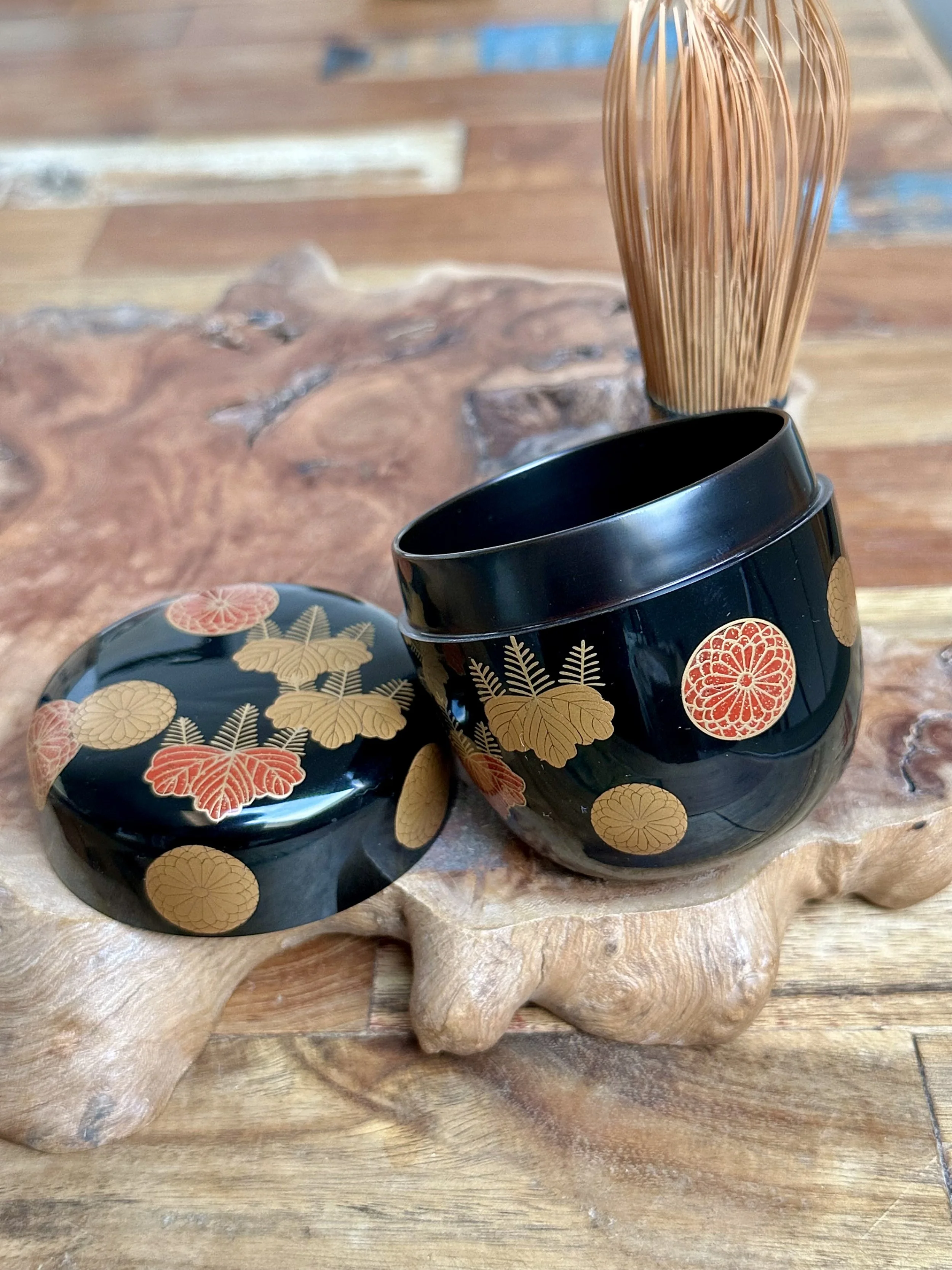 Yūsai lacquered natsume tea caddy, black, with lid and body displaying gold and red chrysanthemum and paulownia crests, next to a bamboo whisk on wood.