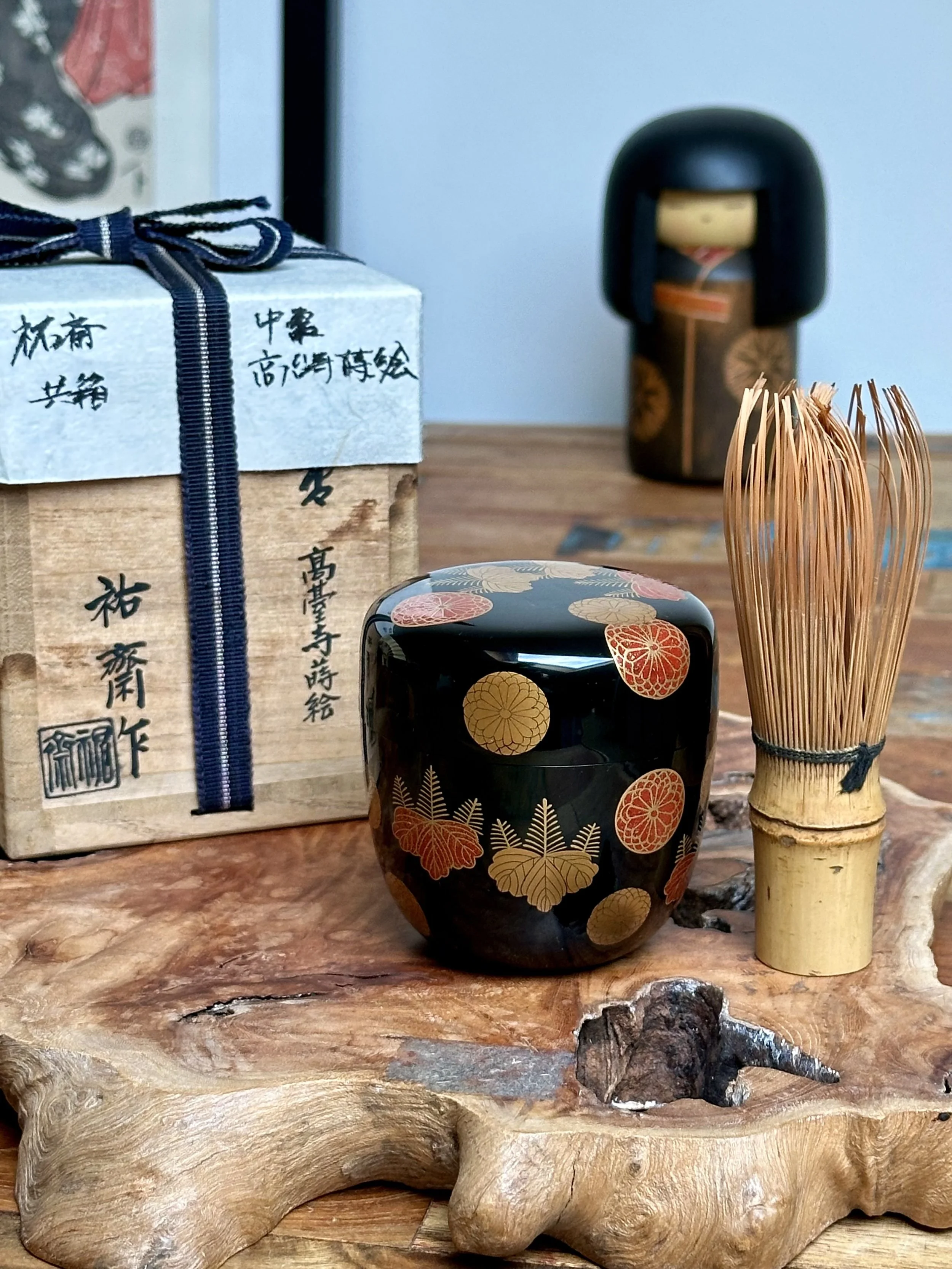 Hariya Yūsai's black lacquered natsume tea caddy with gold and red crests, a bamboo whisk, and its signed tomobako with a blue ribbon, on a rustic wood slab.