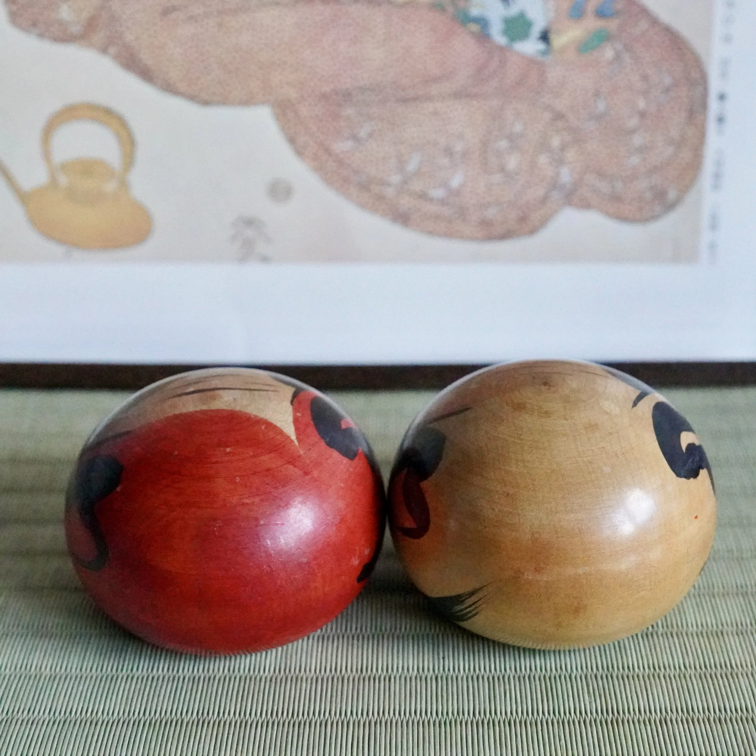 Red and natural wood Tsugaru daruma by Satō Zenji, featuring hand-painted black facial features, resting on a woven mat with blurred artwork behind.