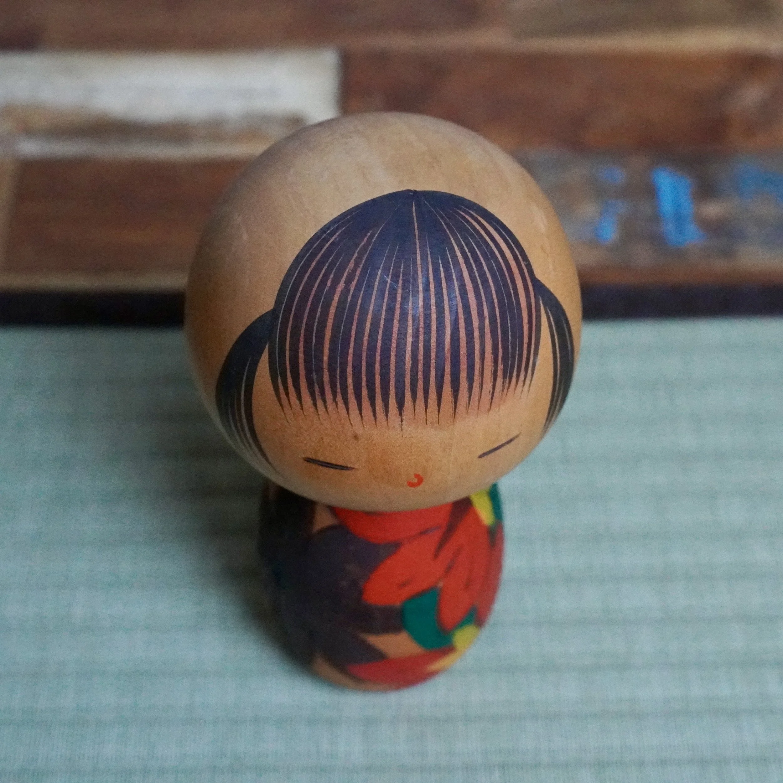 Sōsaku kokeshi by Yuji Kawase, front view. Its light brown head has black painted hair, closed eyes, and a red mouth. The body is carved with red, dark, green, and yellow autumn leaves.